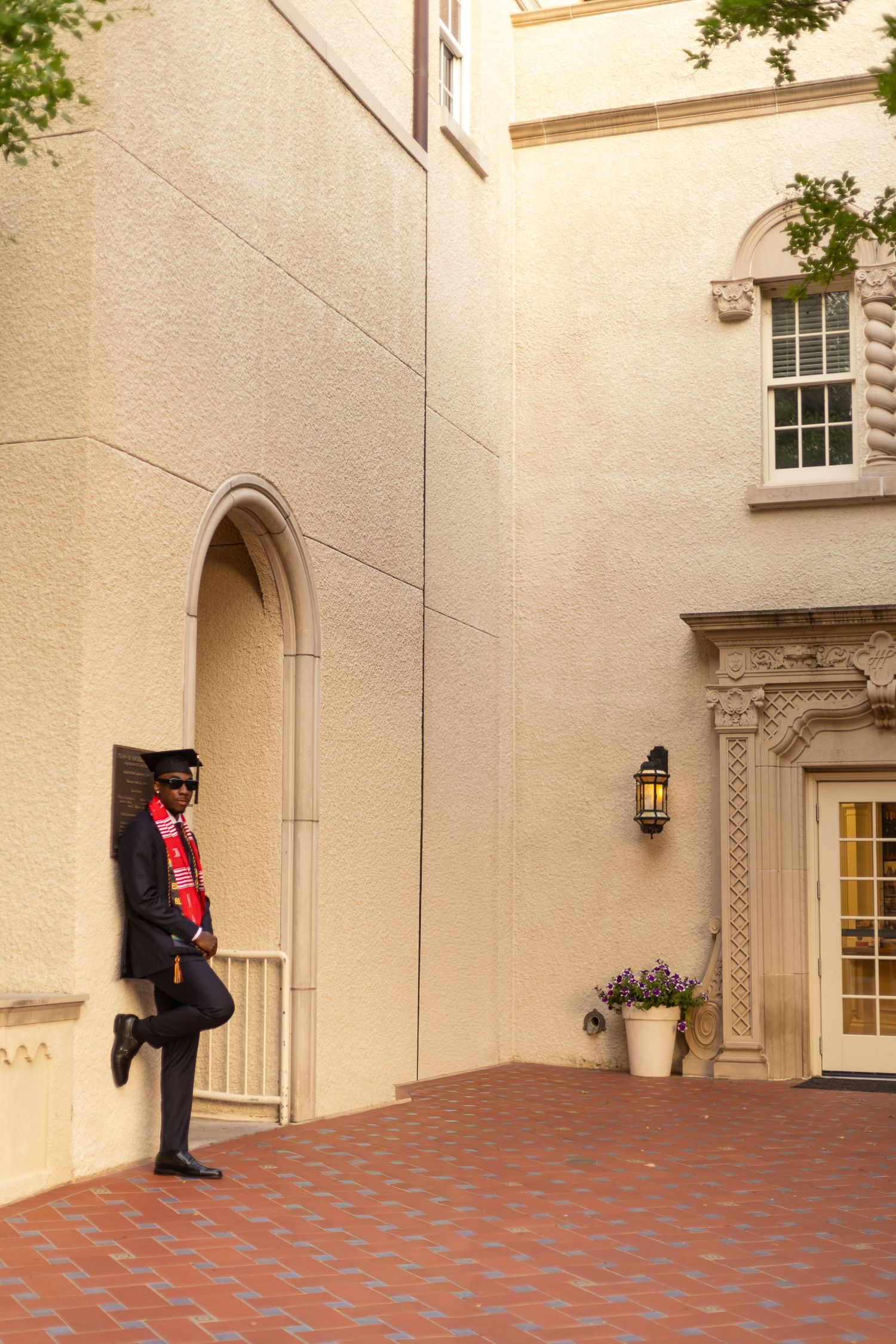 Graduate in cap and gown with red scarf leaning against a beige stone building wall.