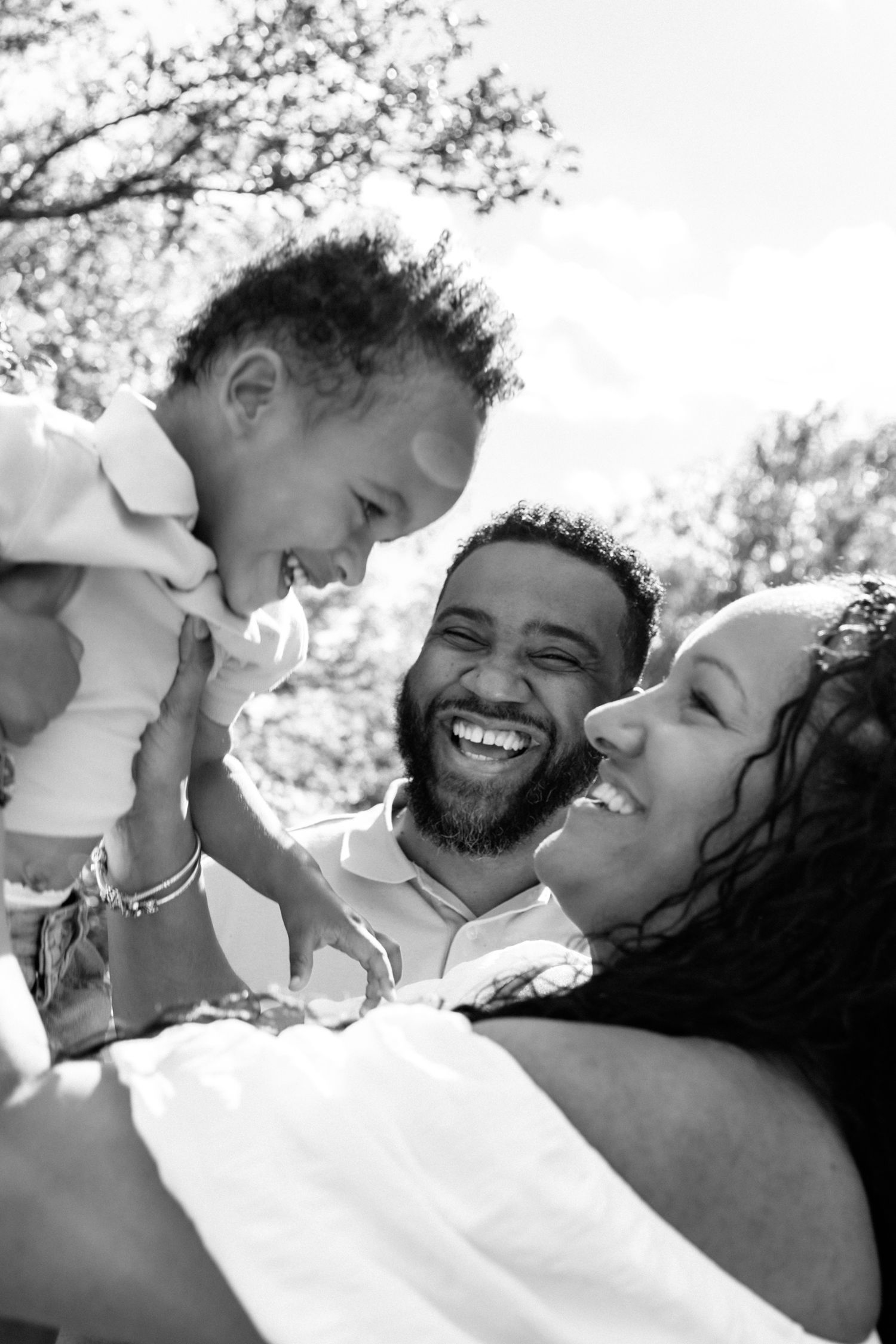 Joyful Black family laughs together as parents lift giggling baby outdoors in black and white photo.
