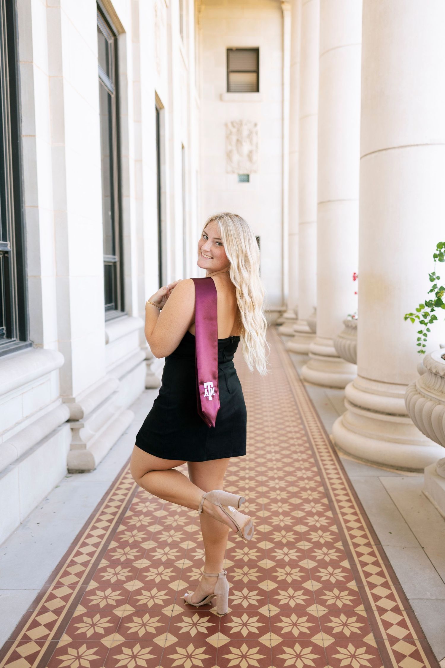 Blonde graduate in black dress with maroon Texas A&M sash posing in elegant columned hallway.