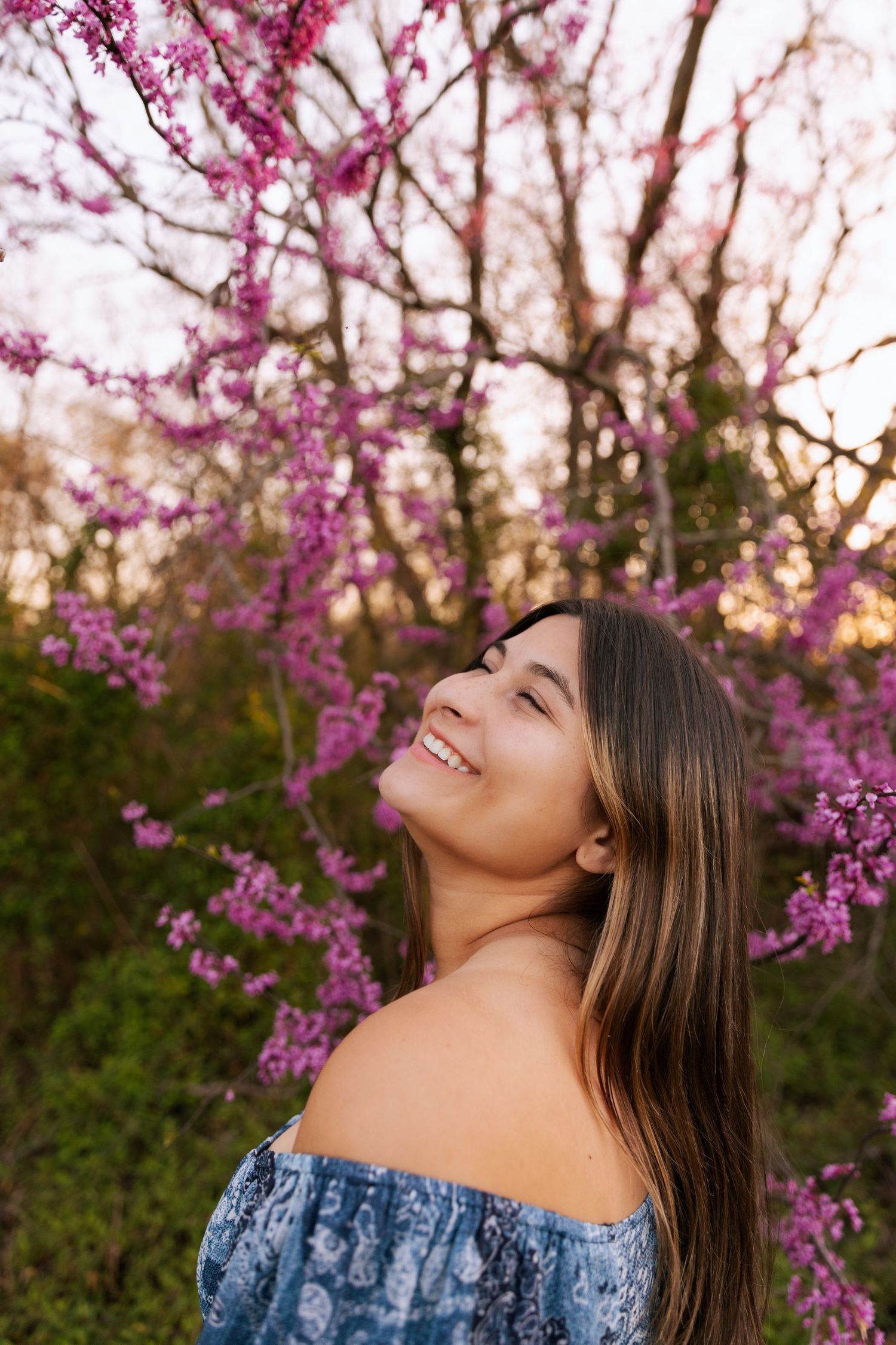 Young woman smiling joyfully among pink cherry blossom trees wearing an off-shoulder blue floral dress.
