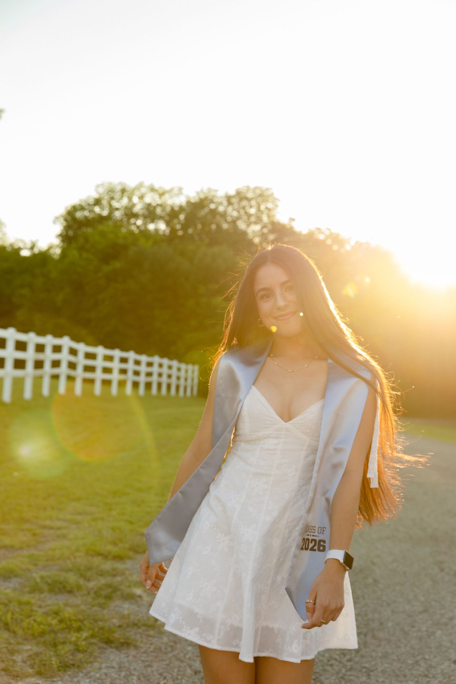 Smiling graduate in white dress and gray gown walks gravel path at golden hour near white fence.