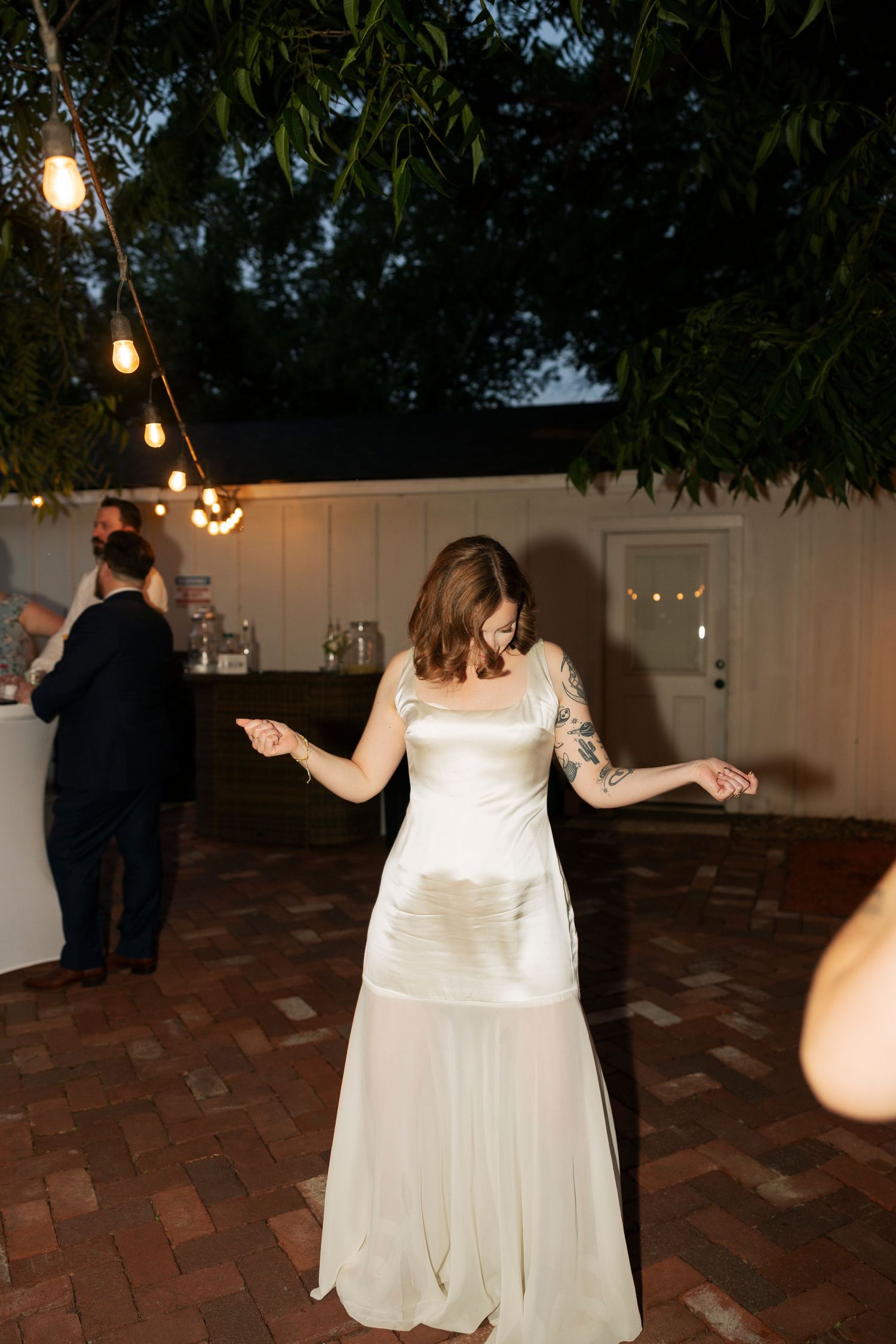 Bride in a white satin gown dancing under string lights at an evening wedding reception.