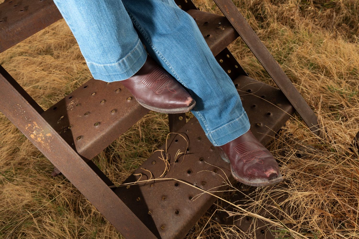 Person wearing brown cowboy boots and blue jeans standing on rusty metal steps surrounded by dry hay.