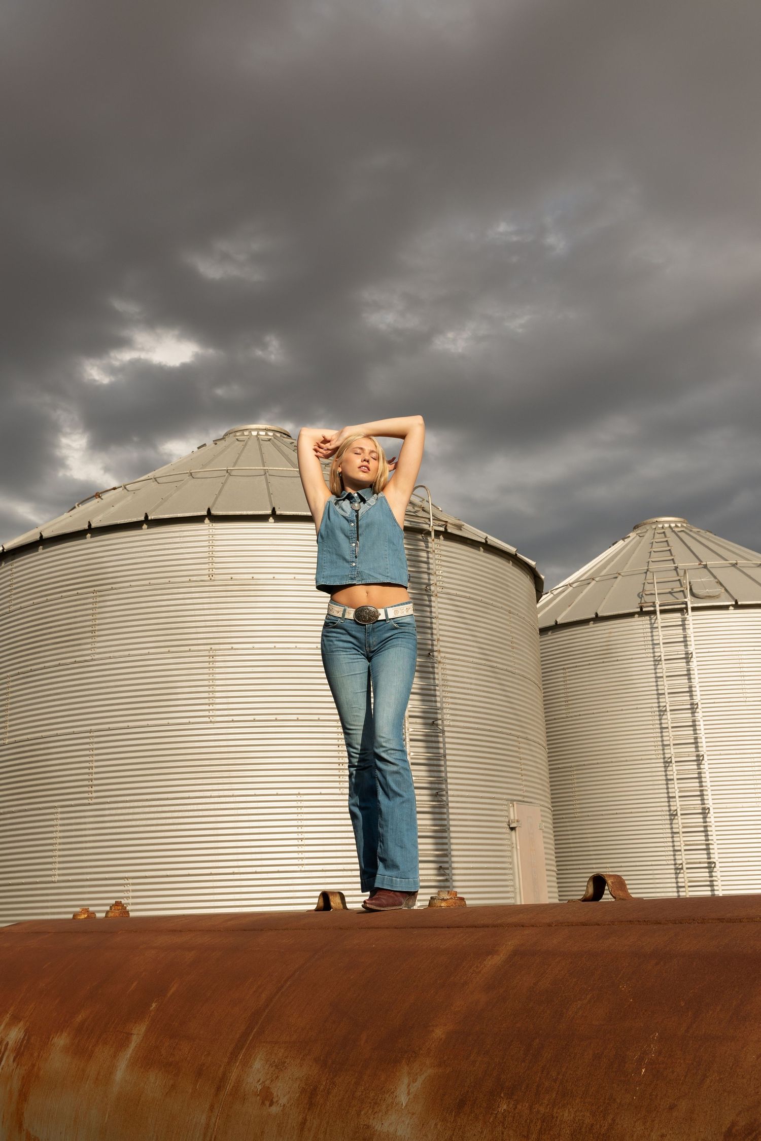 A blonde woman in denim poses atop a rusty tank before two large grain silos under a dramatic stormy sky.