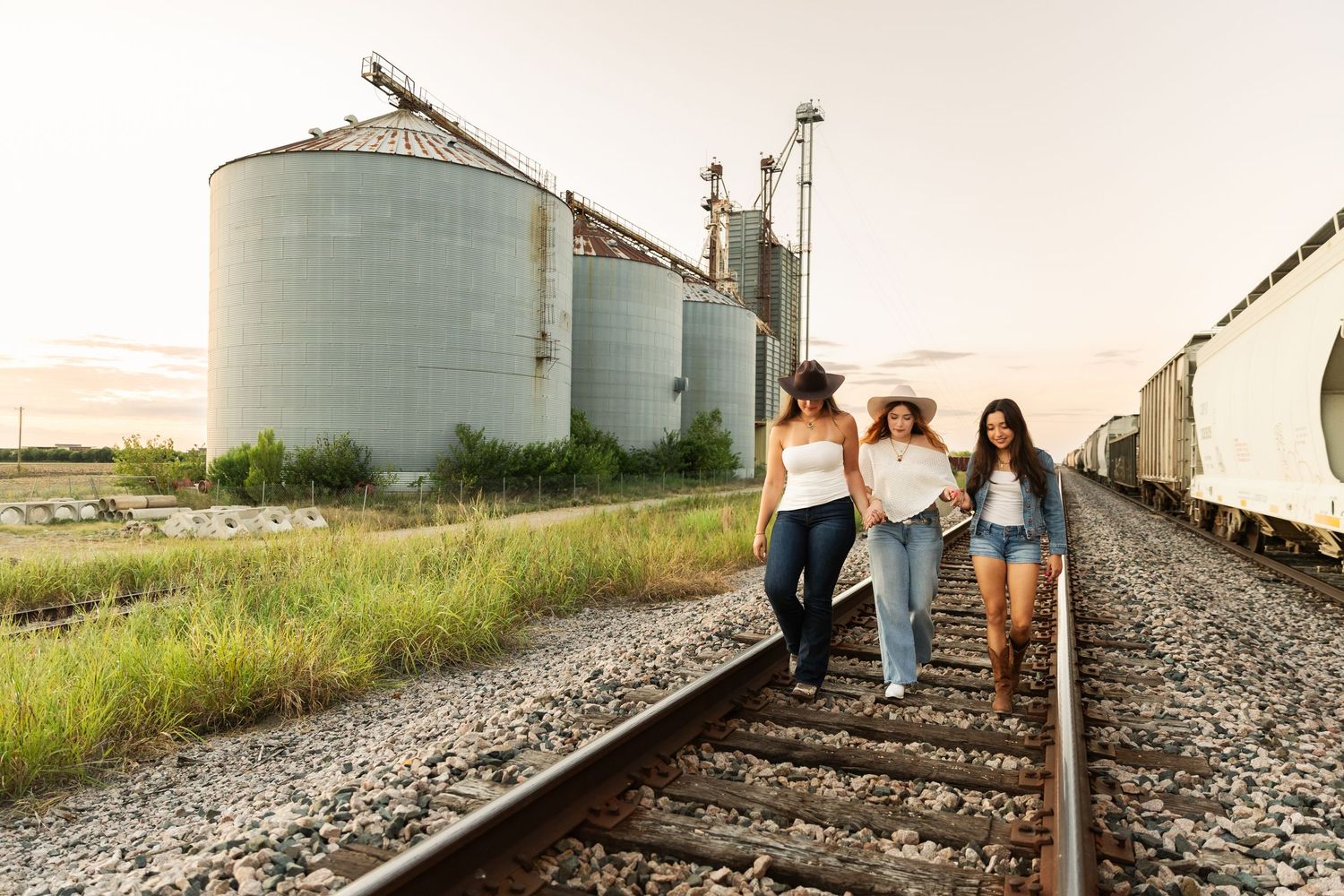Three women in cowboy hats walk along railroad tracks near grain silos at sunset.