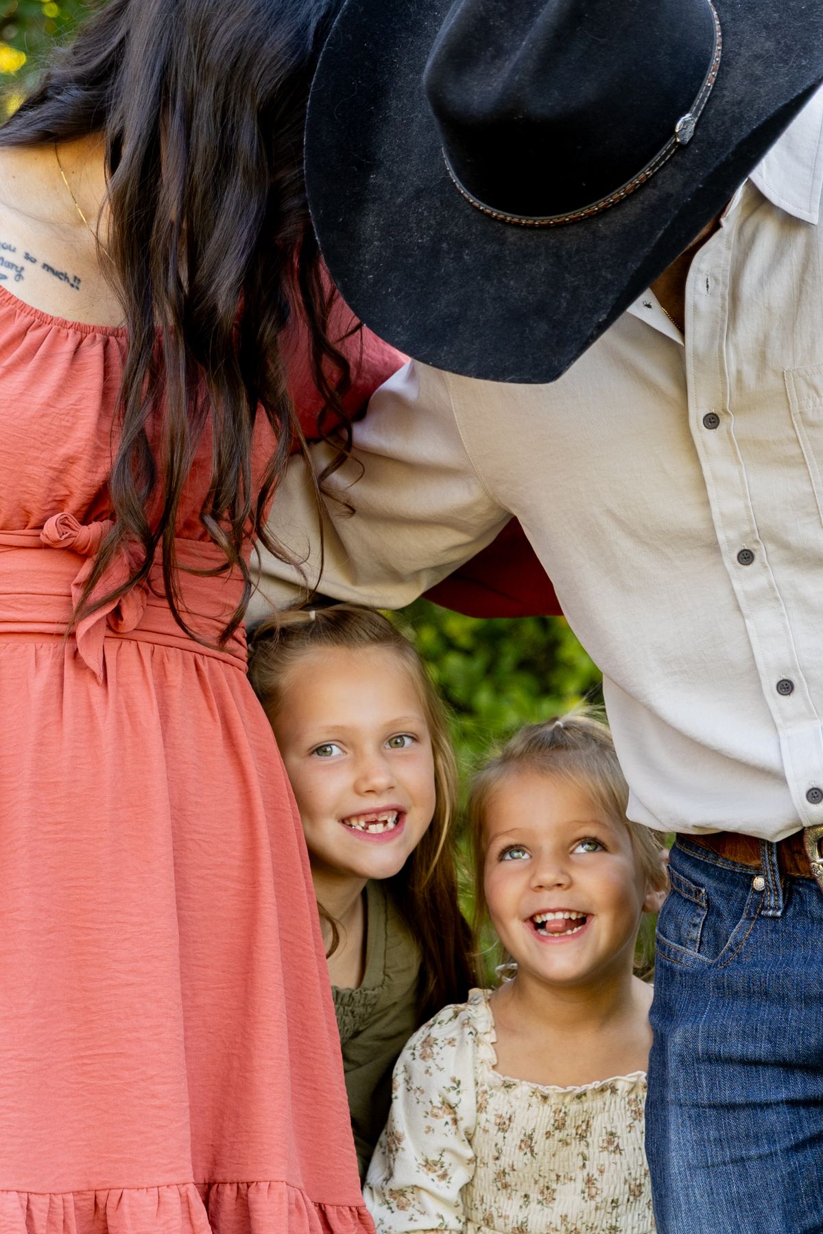 Two smiling young girls peek out between a woman in a coral dress and a man in a black cowboy hat during an outdoor family photo.