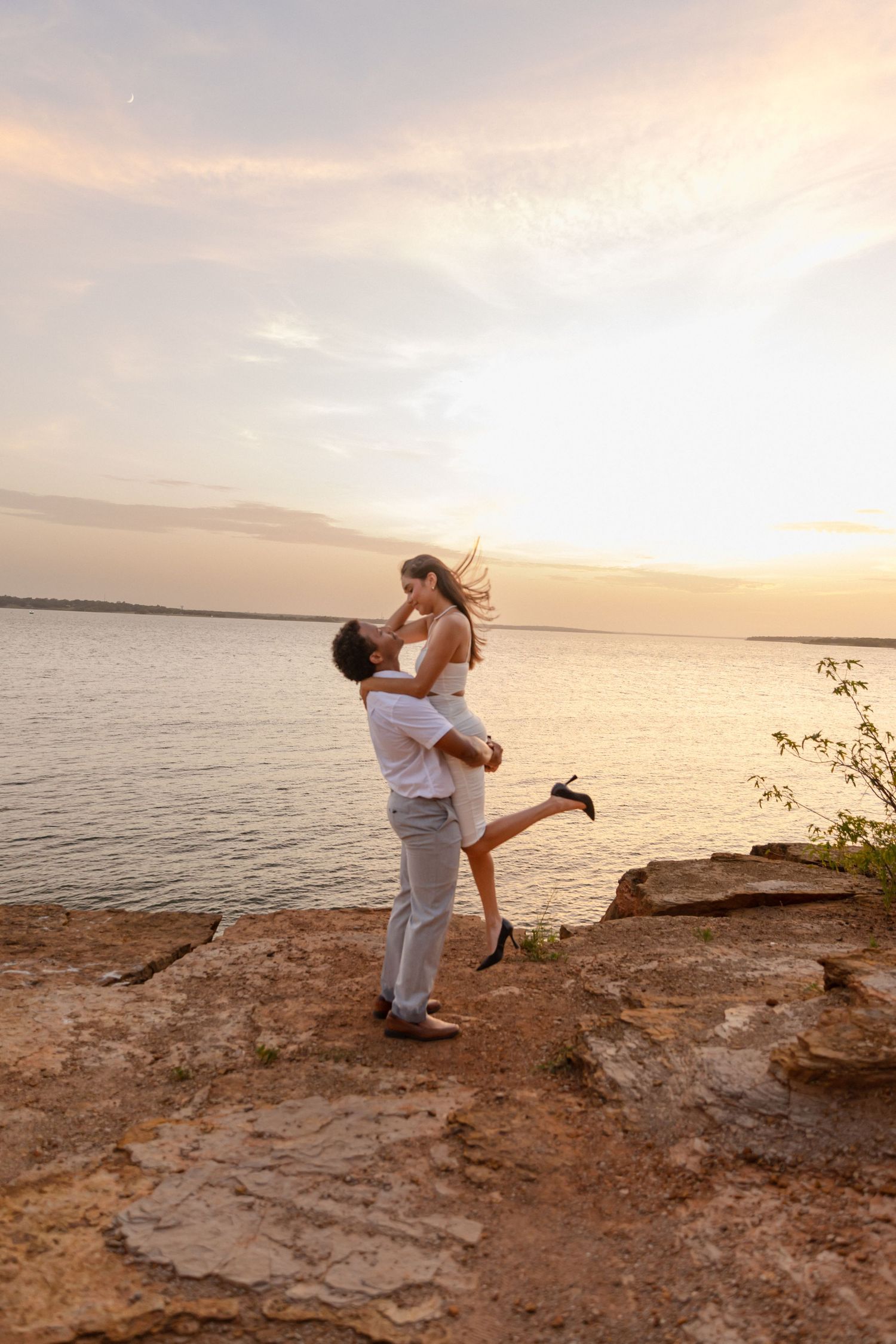 Romantic couple embracing at sunset on rocky shore, man lifting woman in white dress with golden water backdrop.