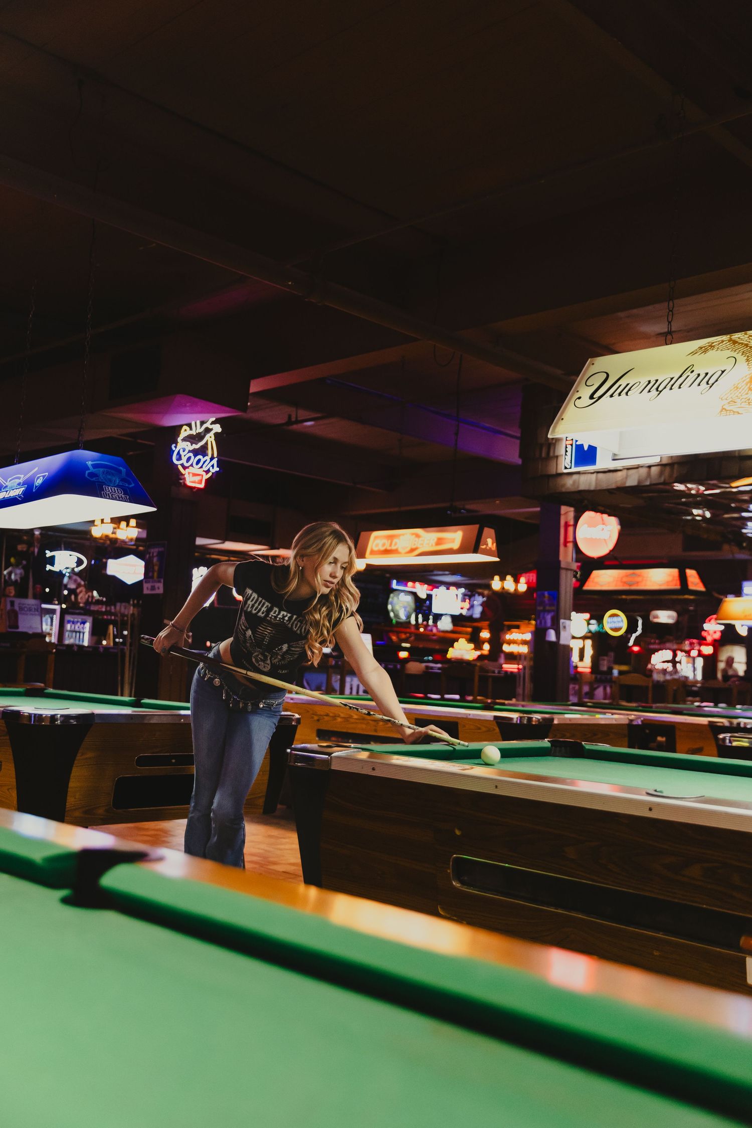 Blonde woman playing pool in a lively bar with neon signs and multiple billiard tables in background.