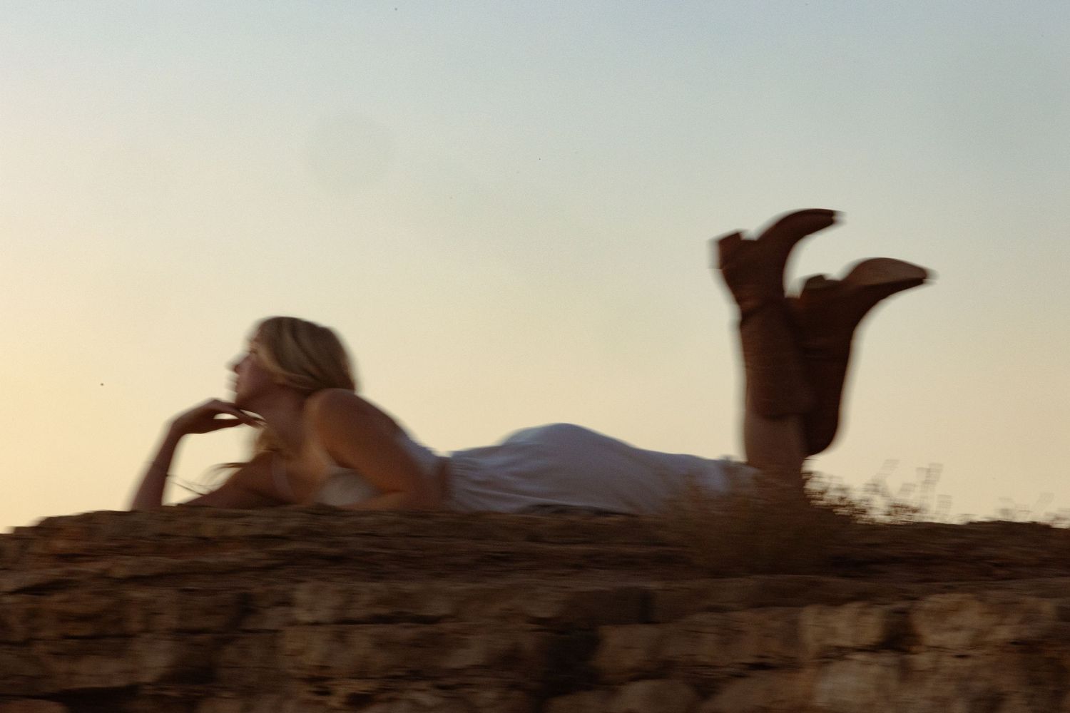 Blonde woman in white dress lying on rocky cliff edge at sunset with legs raised.
