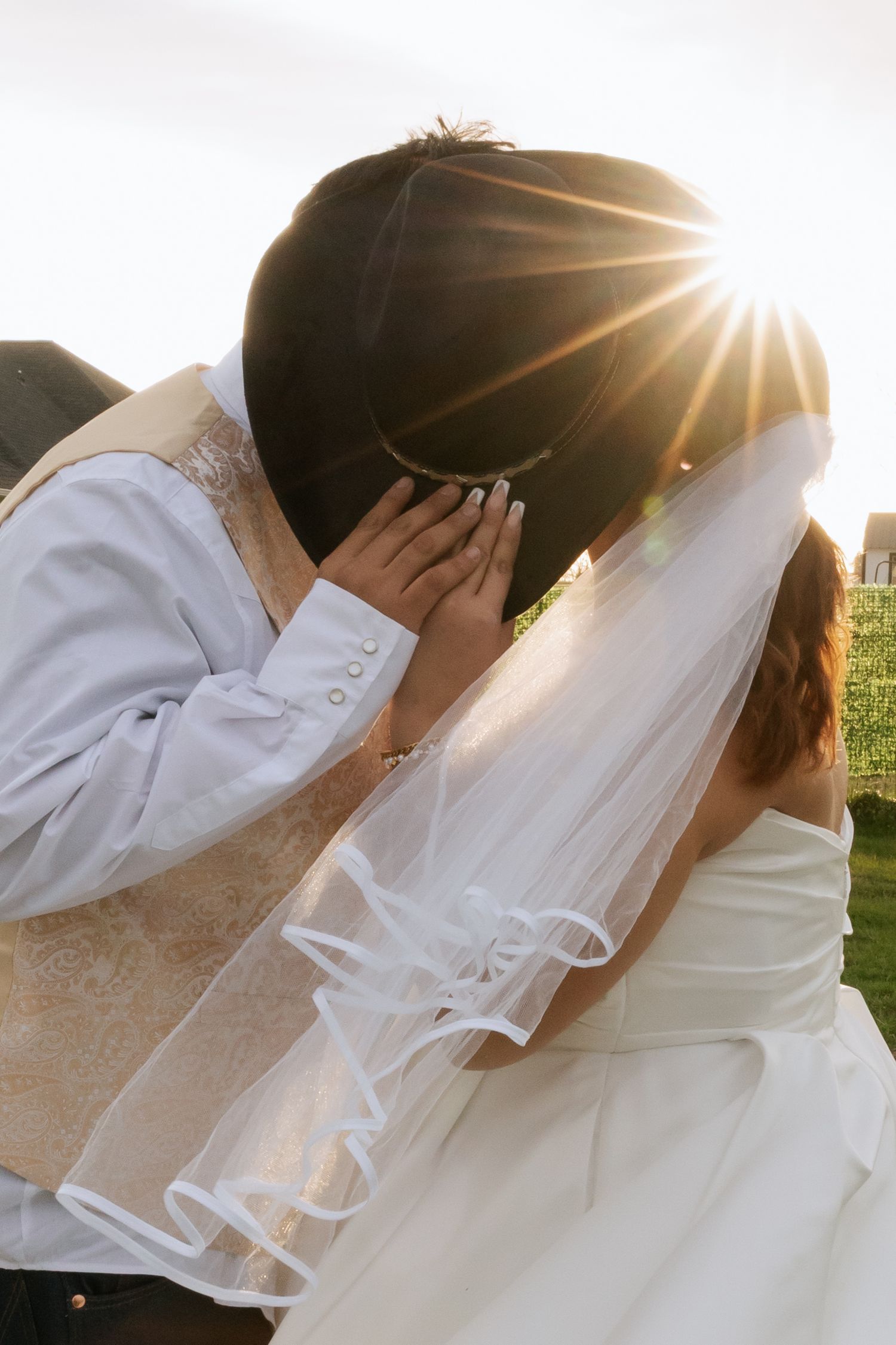 Romantic backlit photo of bride and groom kissing with sun flare bursting through grooms cowboy hat.