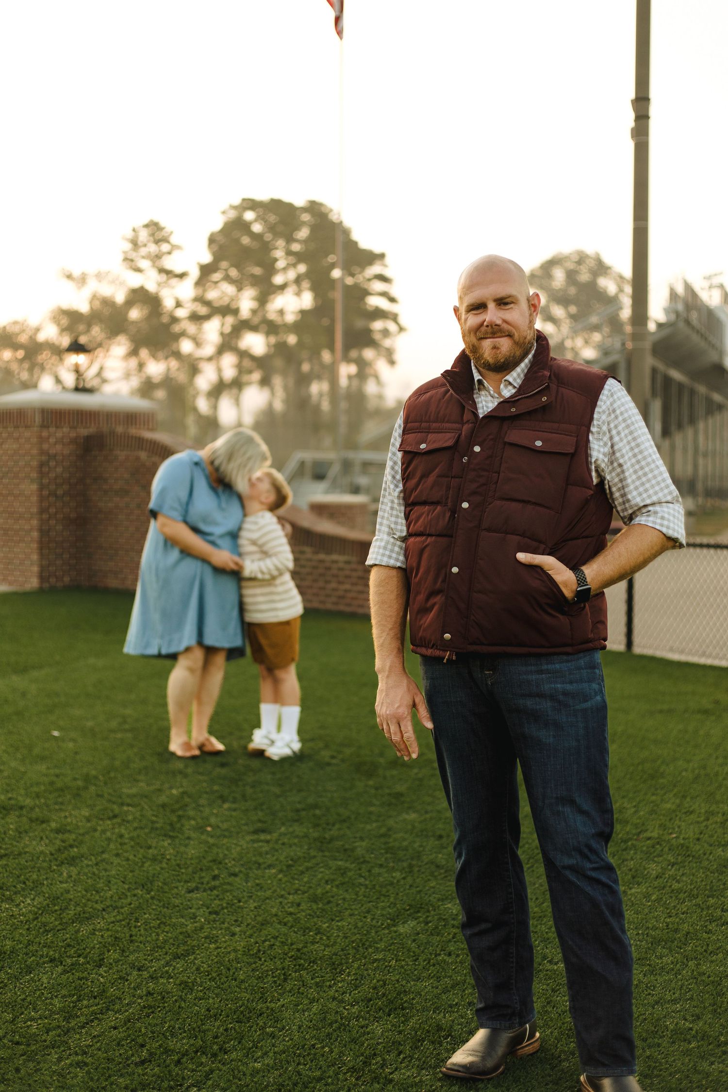 Bald bearded man in maroon vest stands on turf while family hugs in background.