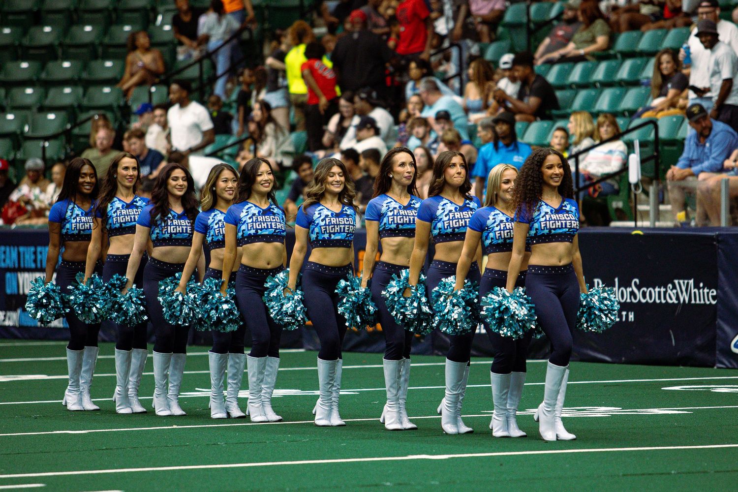 Row of cheerleaders in blue uniforms with pom poms performing at indoor football game.