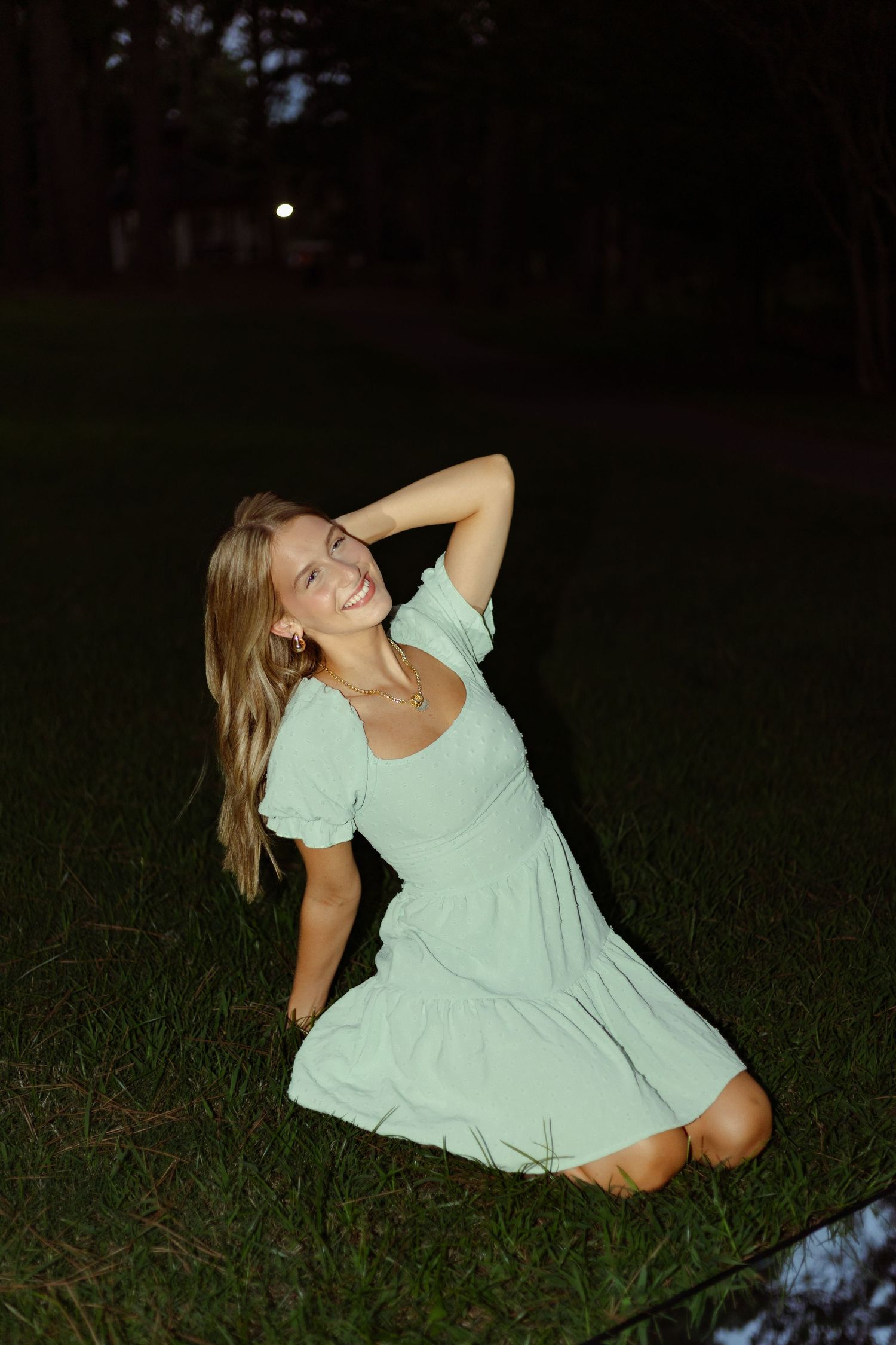 Young blonde woman in a mint green dress smiling while kneeling on grass at night outdoors.