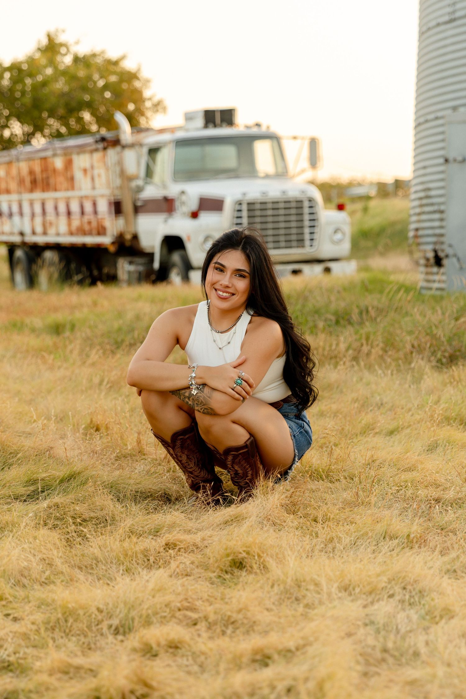 Young woman in cowboy boots and denim shorts crouching in golden field near vintage farm truck at sunset.