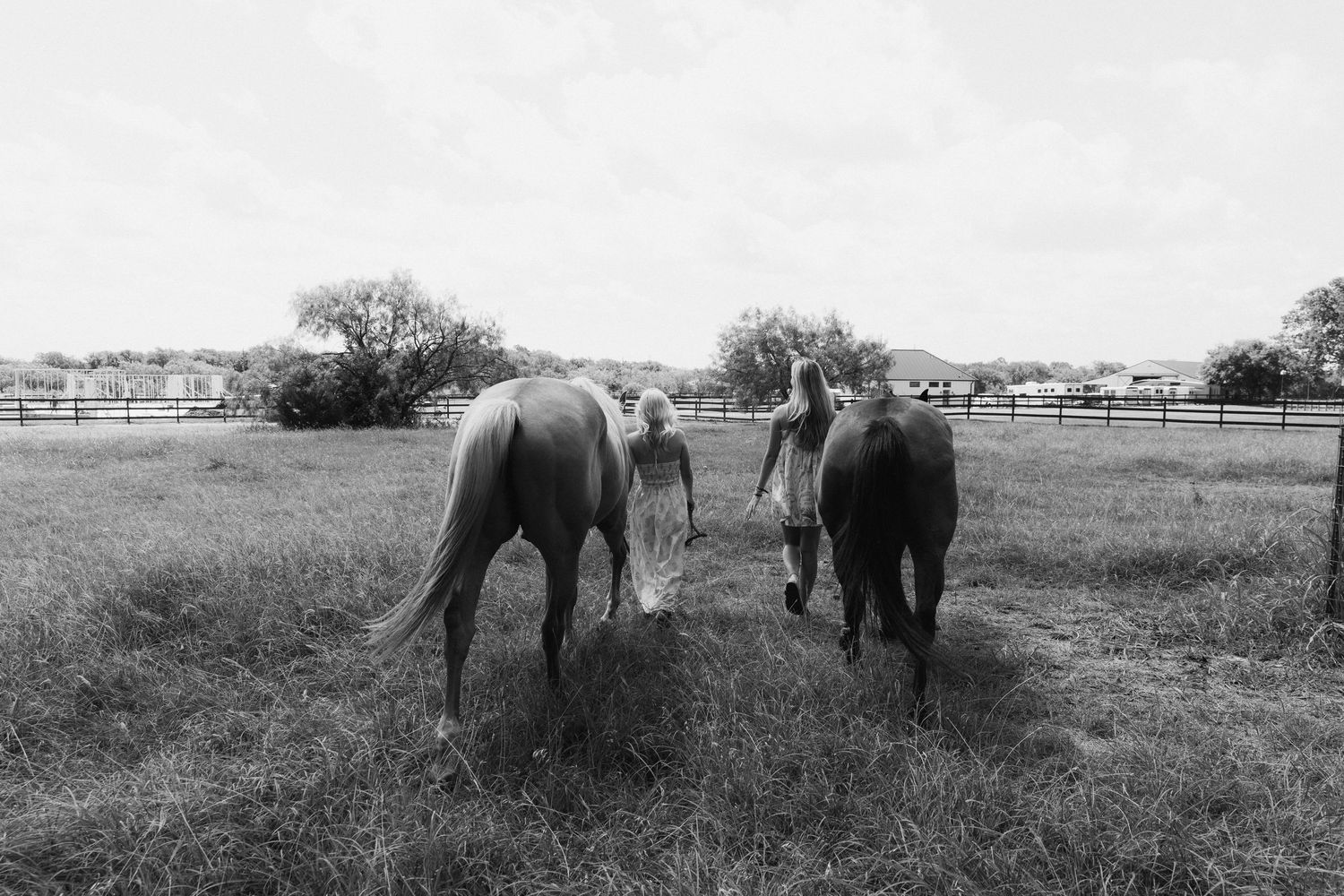 Two women walk with horses through a rustic fenced pasture in a serene black and white countryside photo.