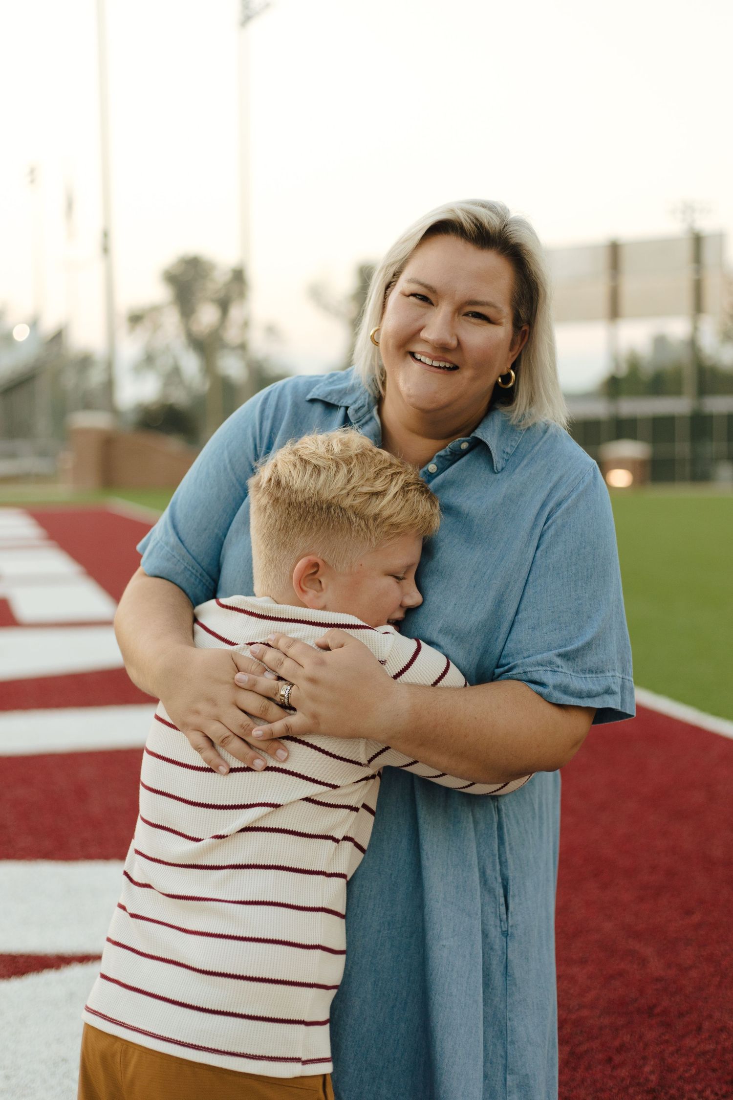 Smiling mother hugging her young blonde son on a sports field at dusk.