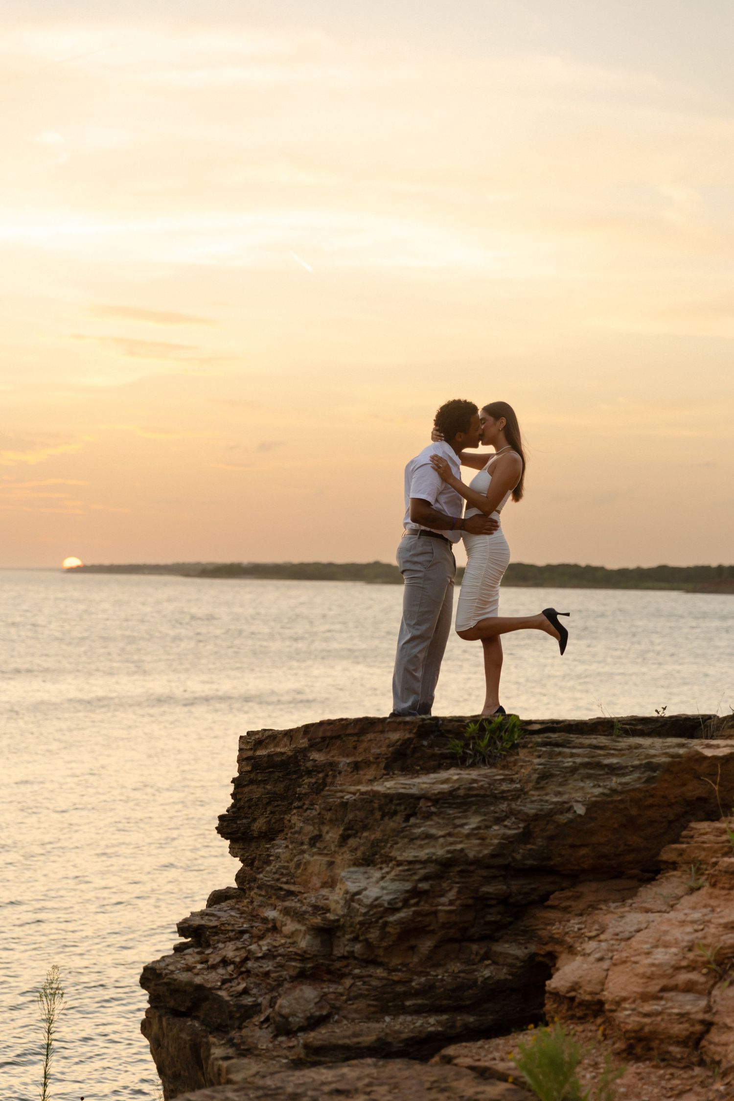 Couple kissing on rocky cliff at sunset with ocean backdrop, romantic silhouette in white outfits.