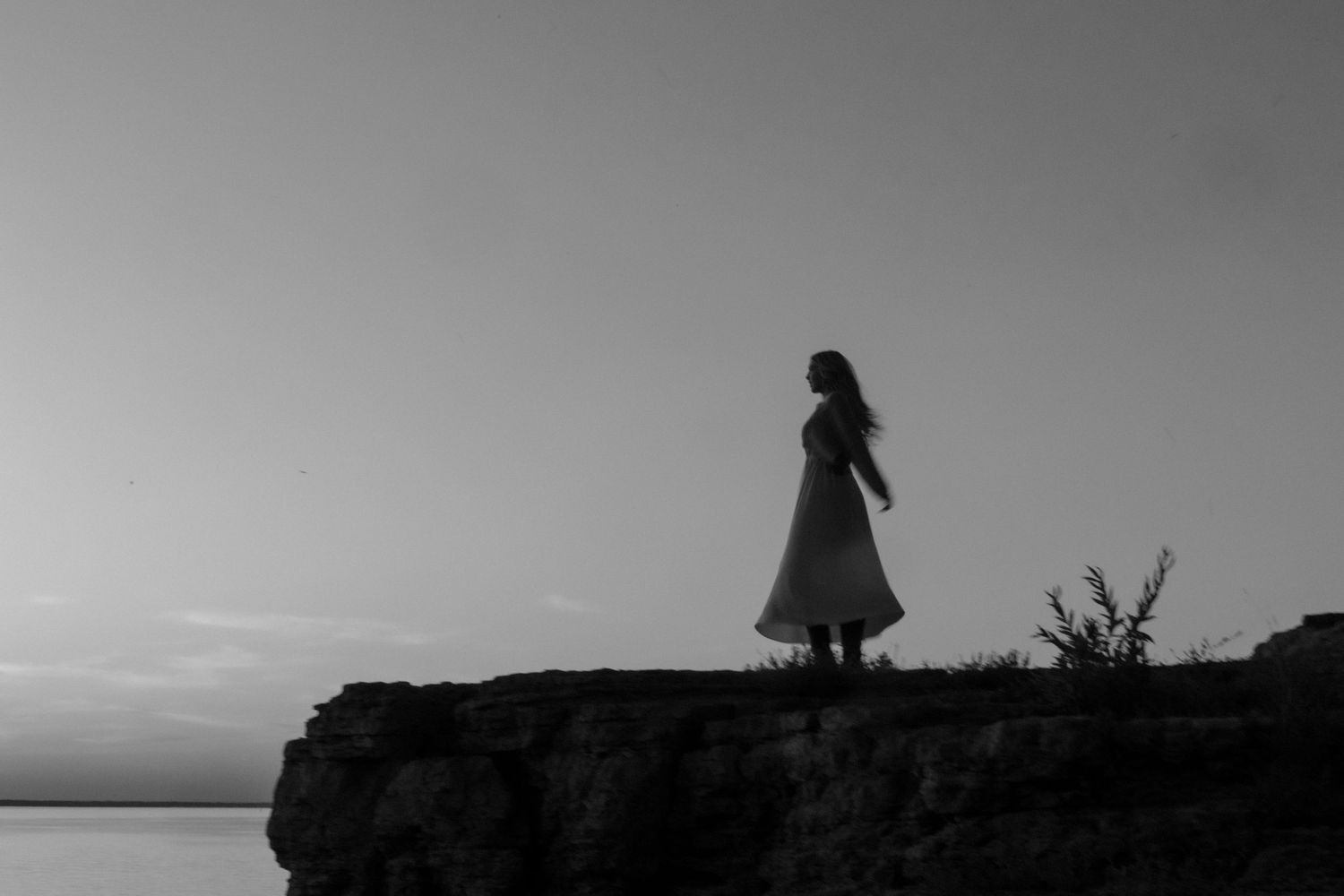 Black and white silhouette of woman in dress standing on rocky cliff overlooking water at dusk.