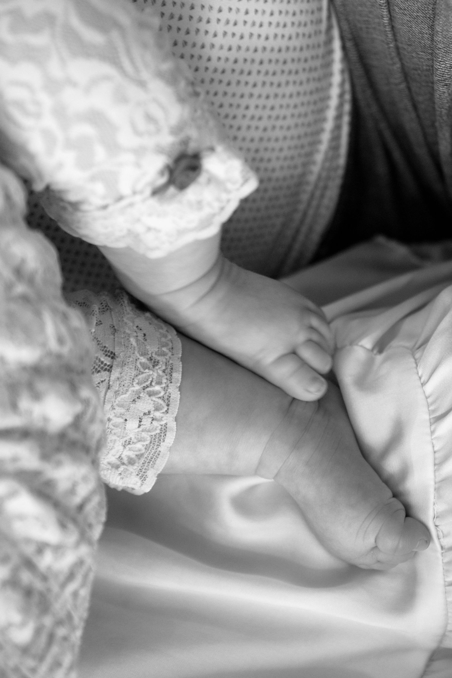Close-up black and white photo of a newborn baby's tiny feet wearing lace socks resting on a satin blanket.