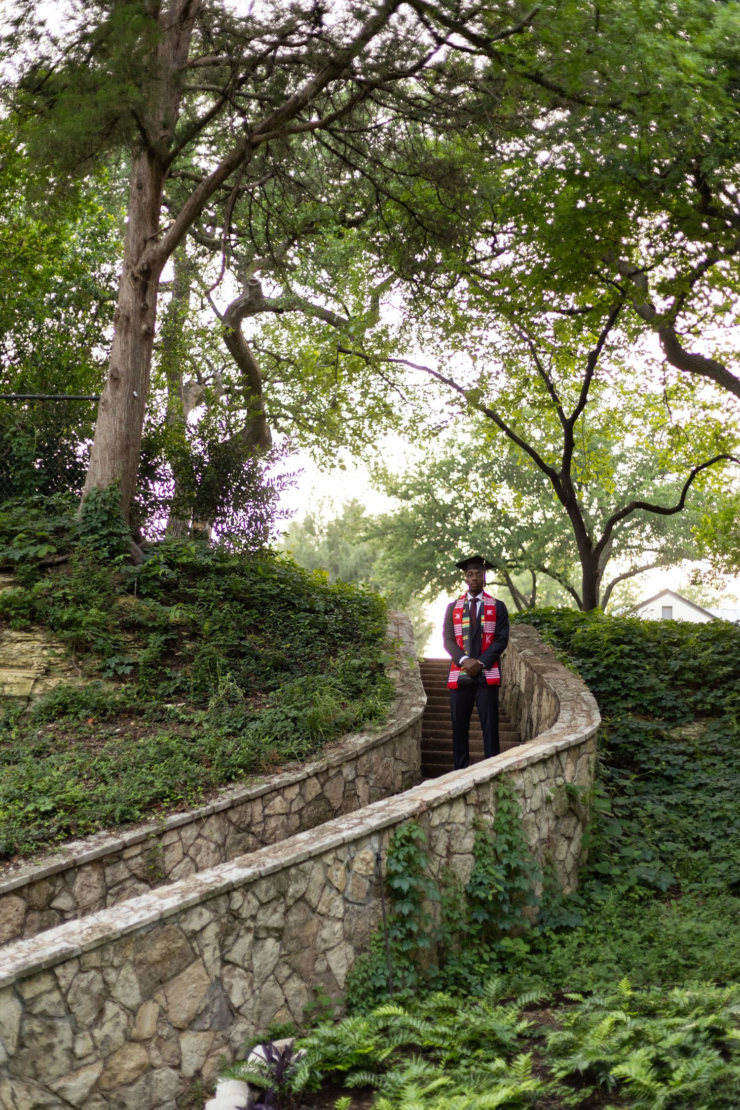 Graduate in cap and gown standing on curved stone wall path surrounded by lush green trees and foliage.