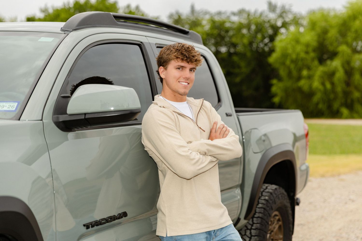 Young man with crossed arms smiling beside a Toyota Tacoma truck outdoors.