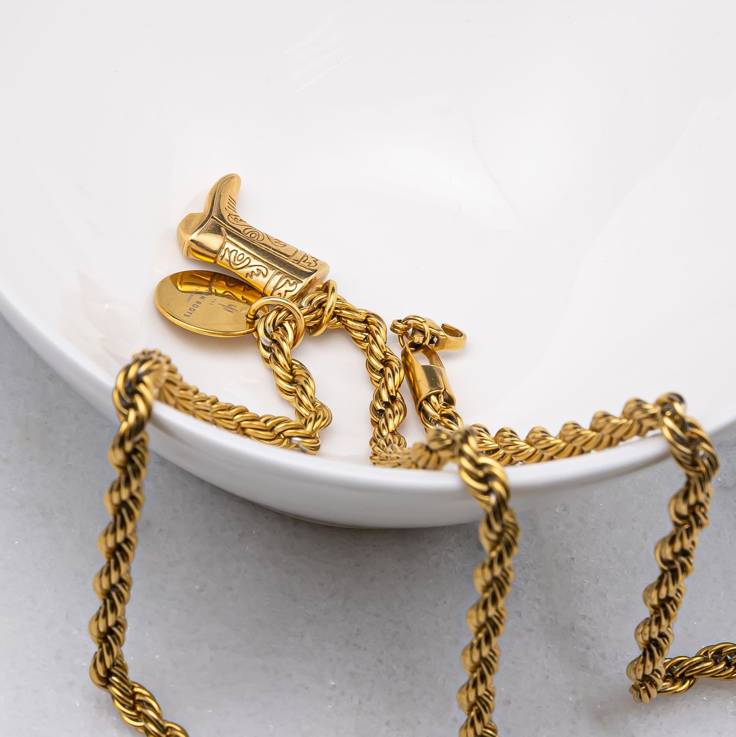Gold rope chain necklace with ornate pendant displayed on white ceramic dish, showcasing luxury jewelry design.