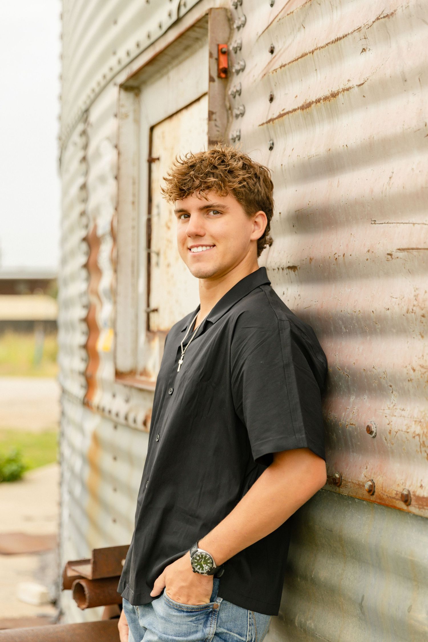Young man in black shirt and jeans smiling while leaning against a rustic corrugated metal wall outdoors.