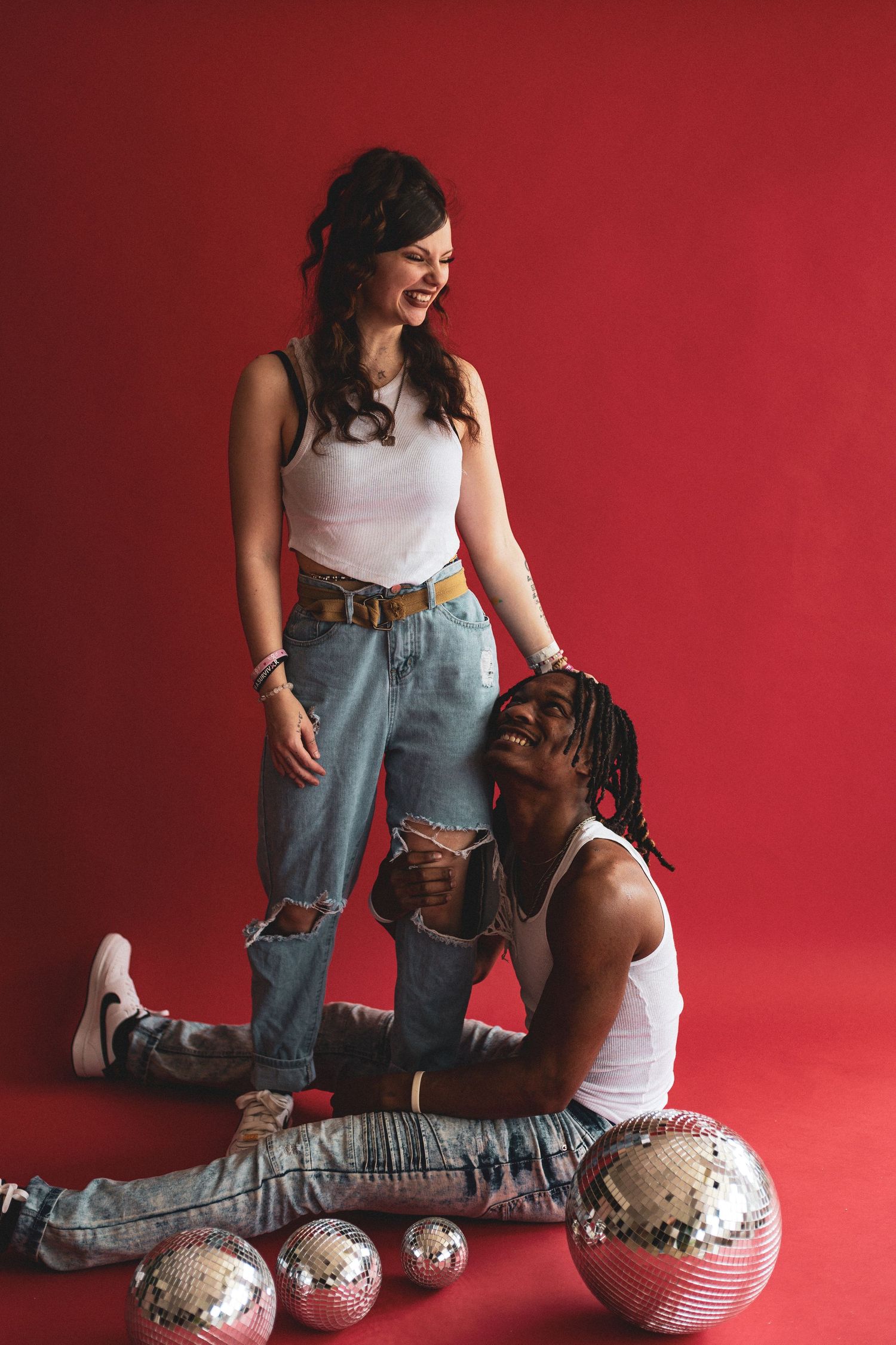 Laughing couple in casual denim outfits posing with disco balls against vibrant red studio background.