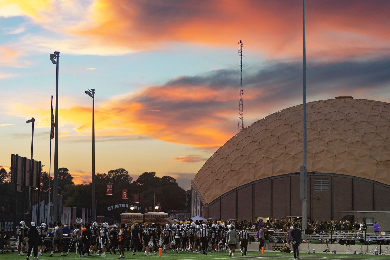 Football teams warming up on field at sunset with dramatic orange sky and domed stadium in background.