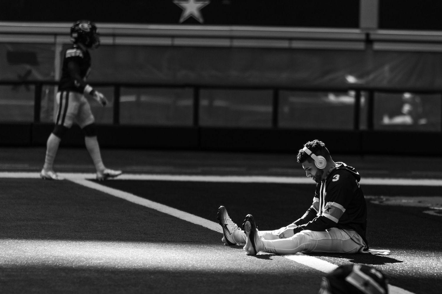 Black and white photo of a football player sitting on the field sideline wearing headphones during a game.