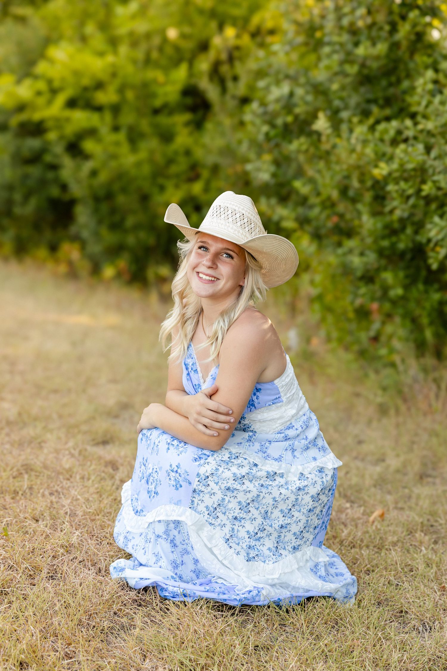 Smiling blonde woman in blue floral dress and straw cowboy hat sitting outdoors near green trees.