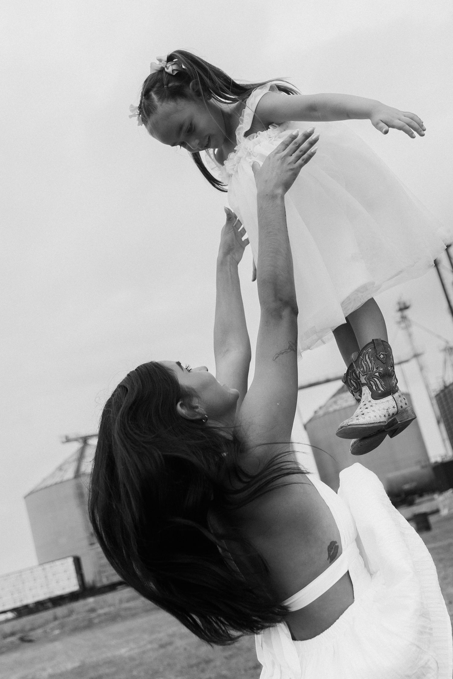 Black and white photo of a woman lifting a young girl in a white dress and cowboy boots into the air outdoors.