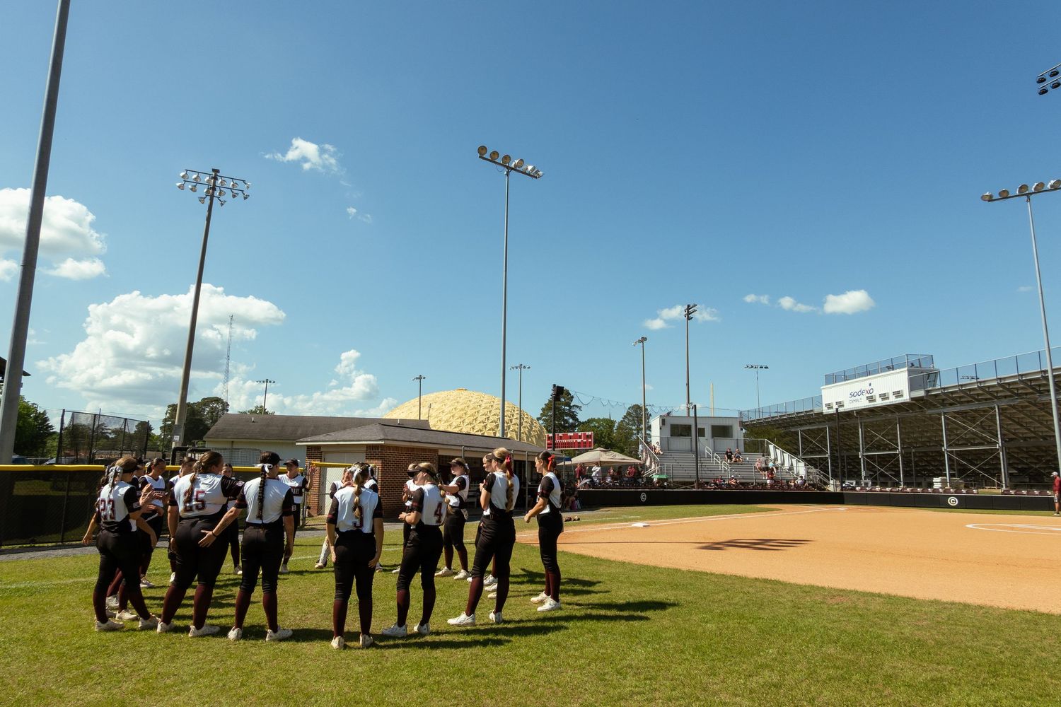 Softball team gathered on field during sunny day at a stadium with lights.