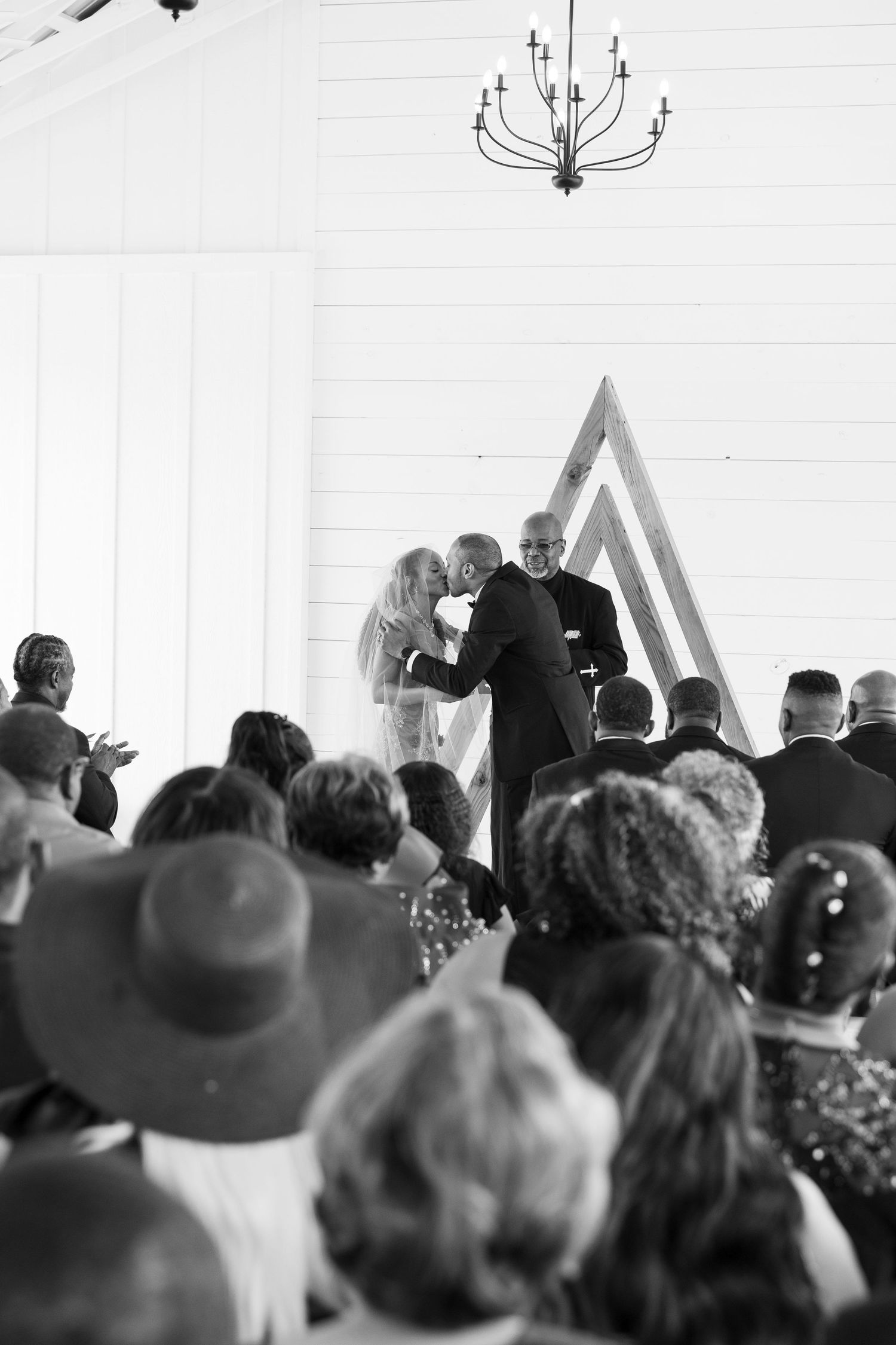 A black and white wide shot of a wedding ceremony inside a white barn with guests seated.