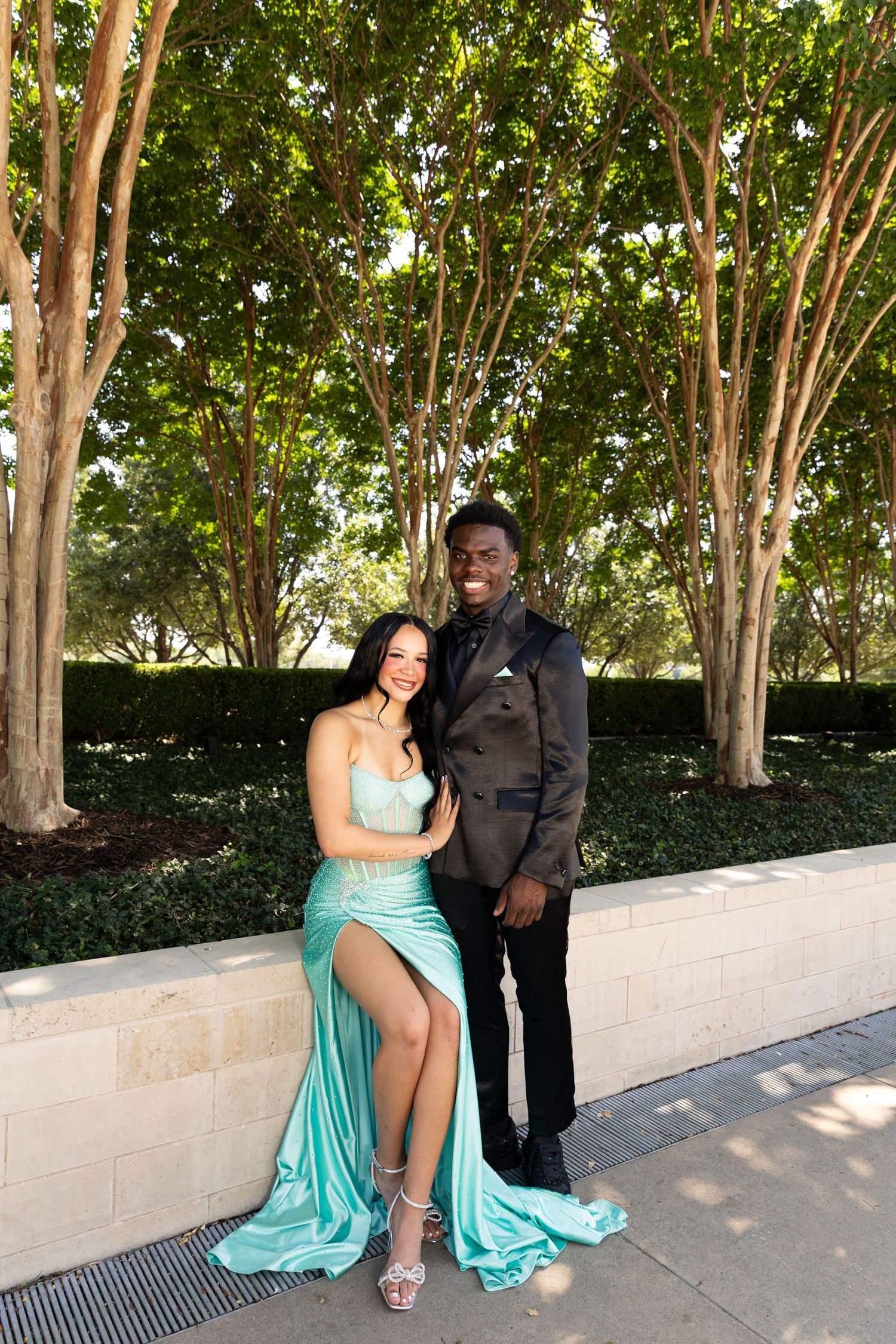 Couple posing outdoors in prom attire, woman in teal satin gown and man in black suit.