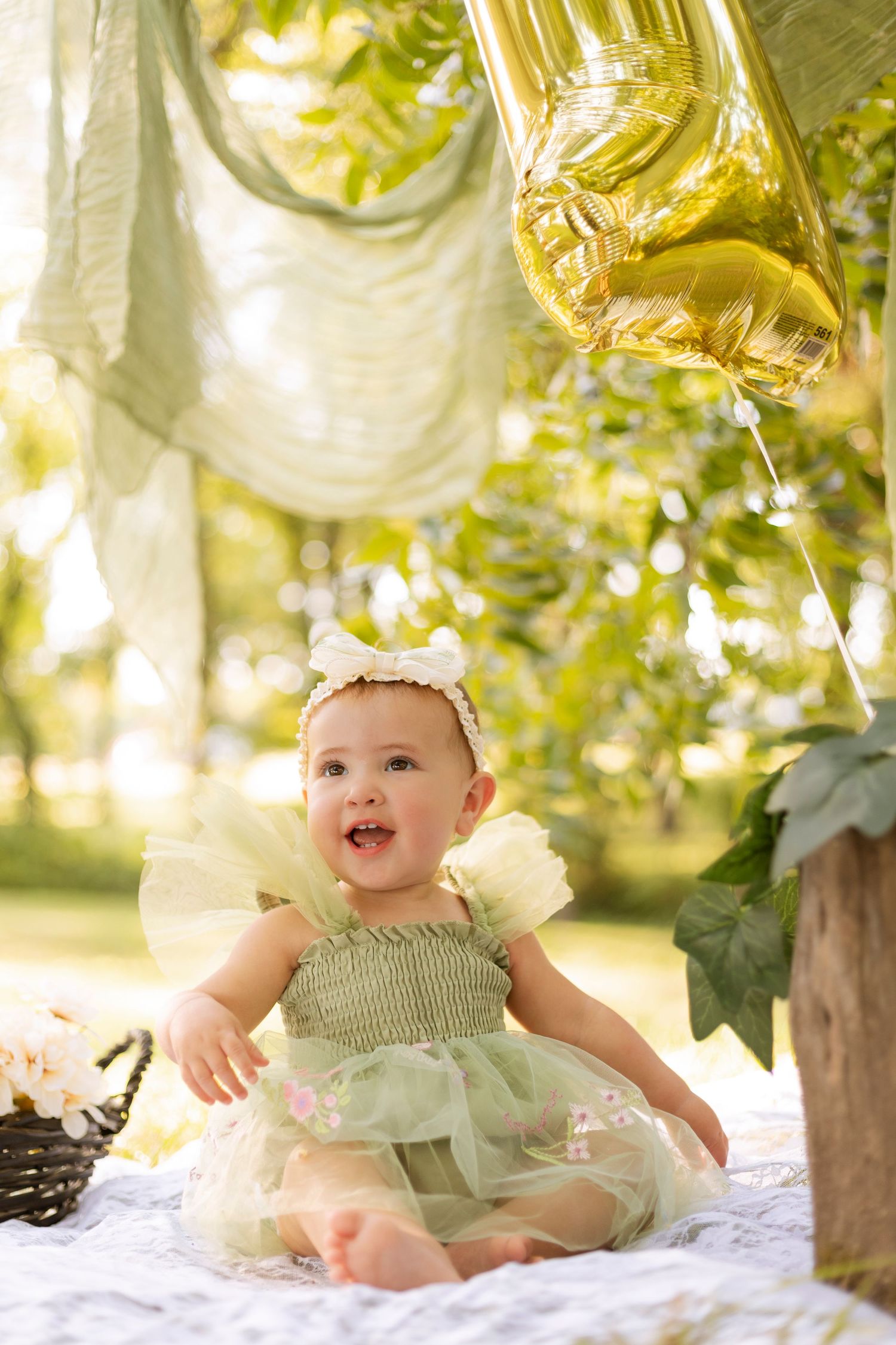 Adorable baby girl in green tutu dress sits outdoors near gold number balloon at birthday photoshoot.
