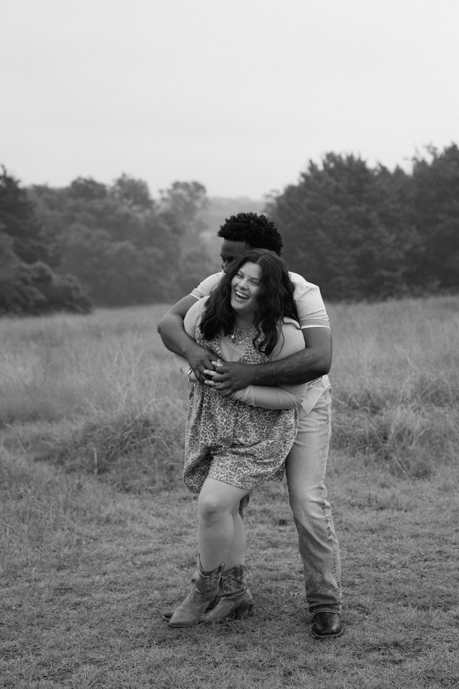 Black and white photo of a laughing couple embracing in an open field with trees in the background.