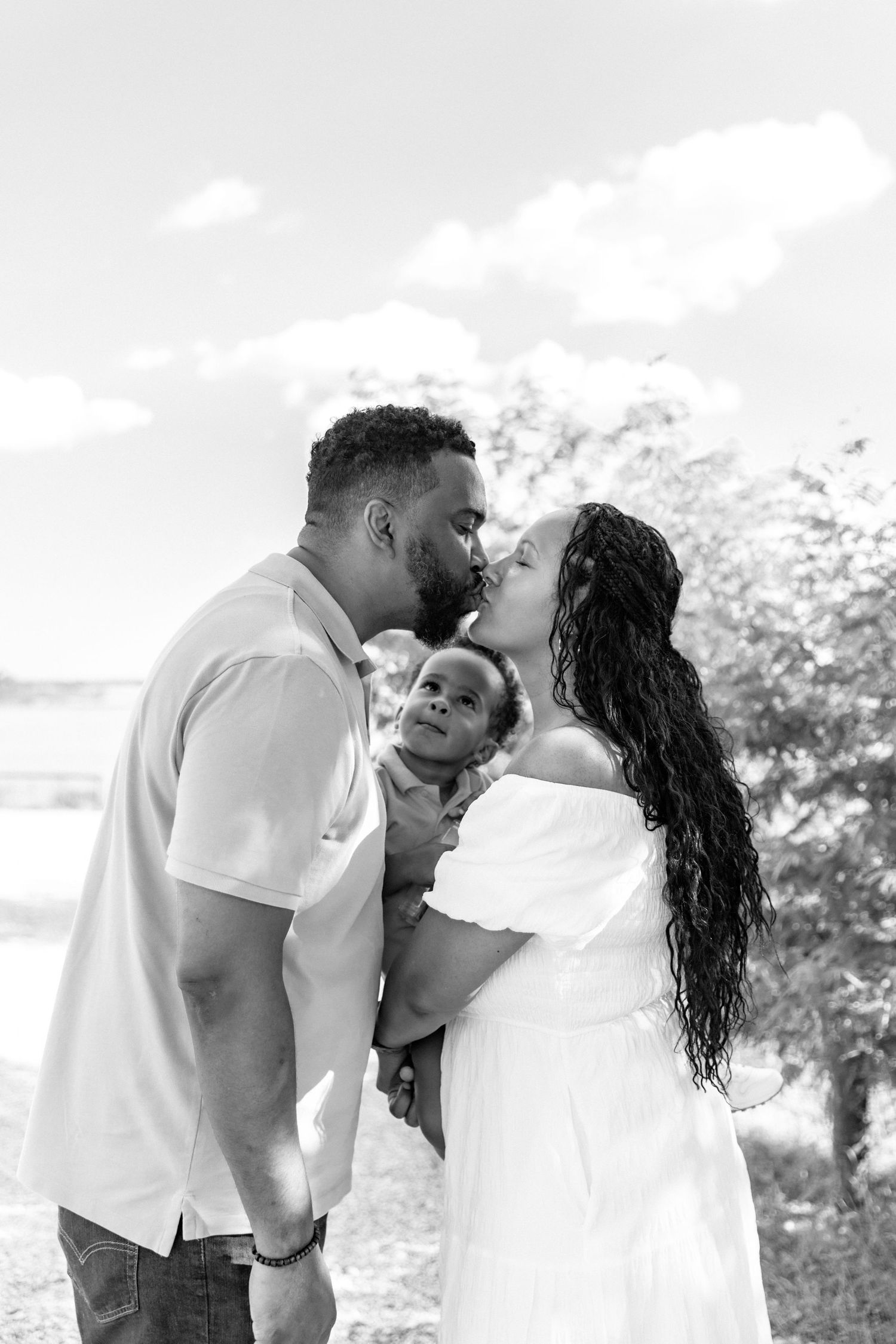 Black and white photo of a couple kissing while holding their baby outdoors near a lake.