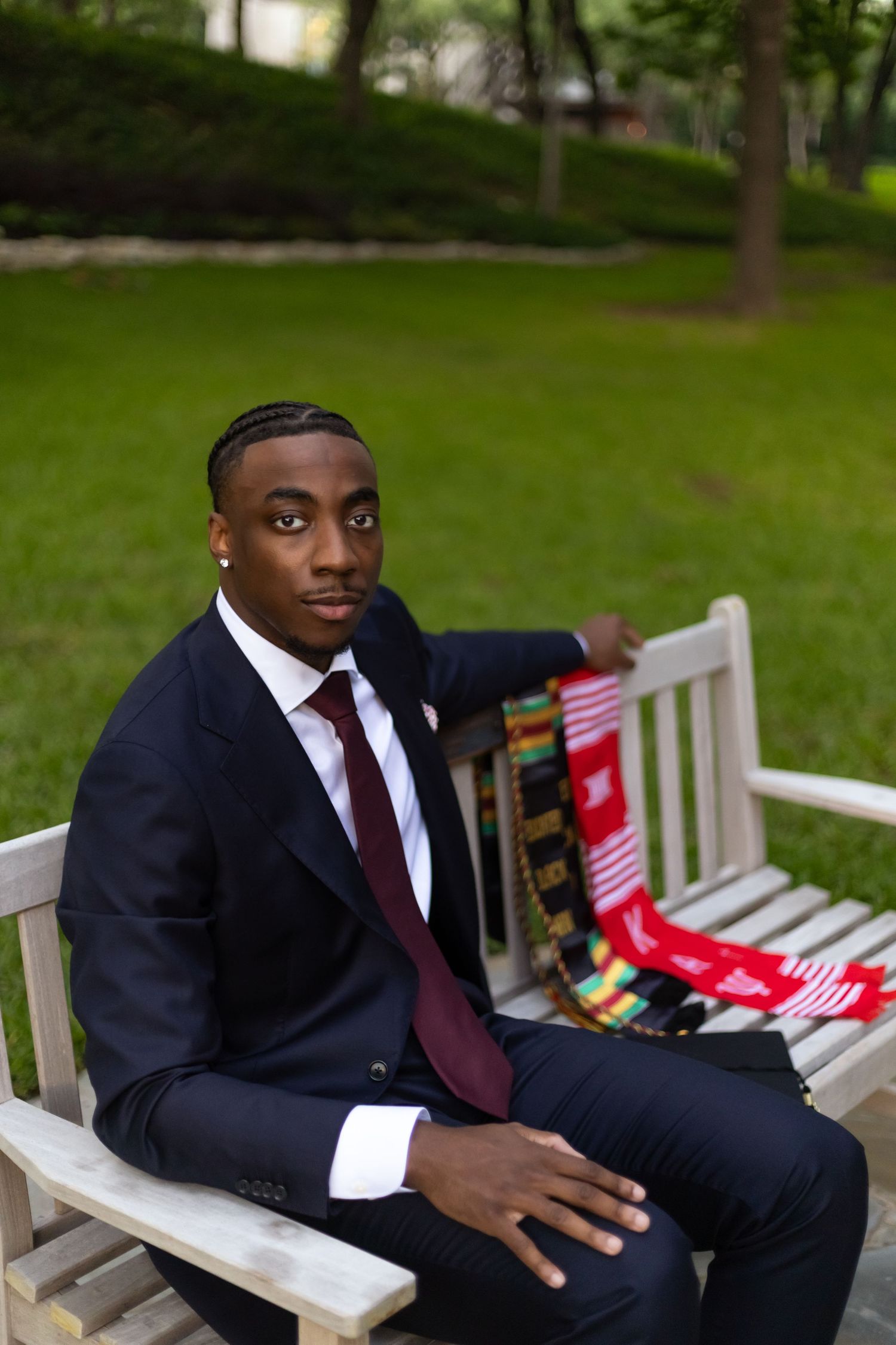 Young man in navy suit with burgundy tie sitting on a white bench in a park.