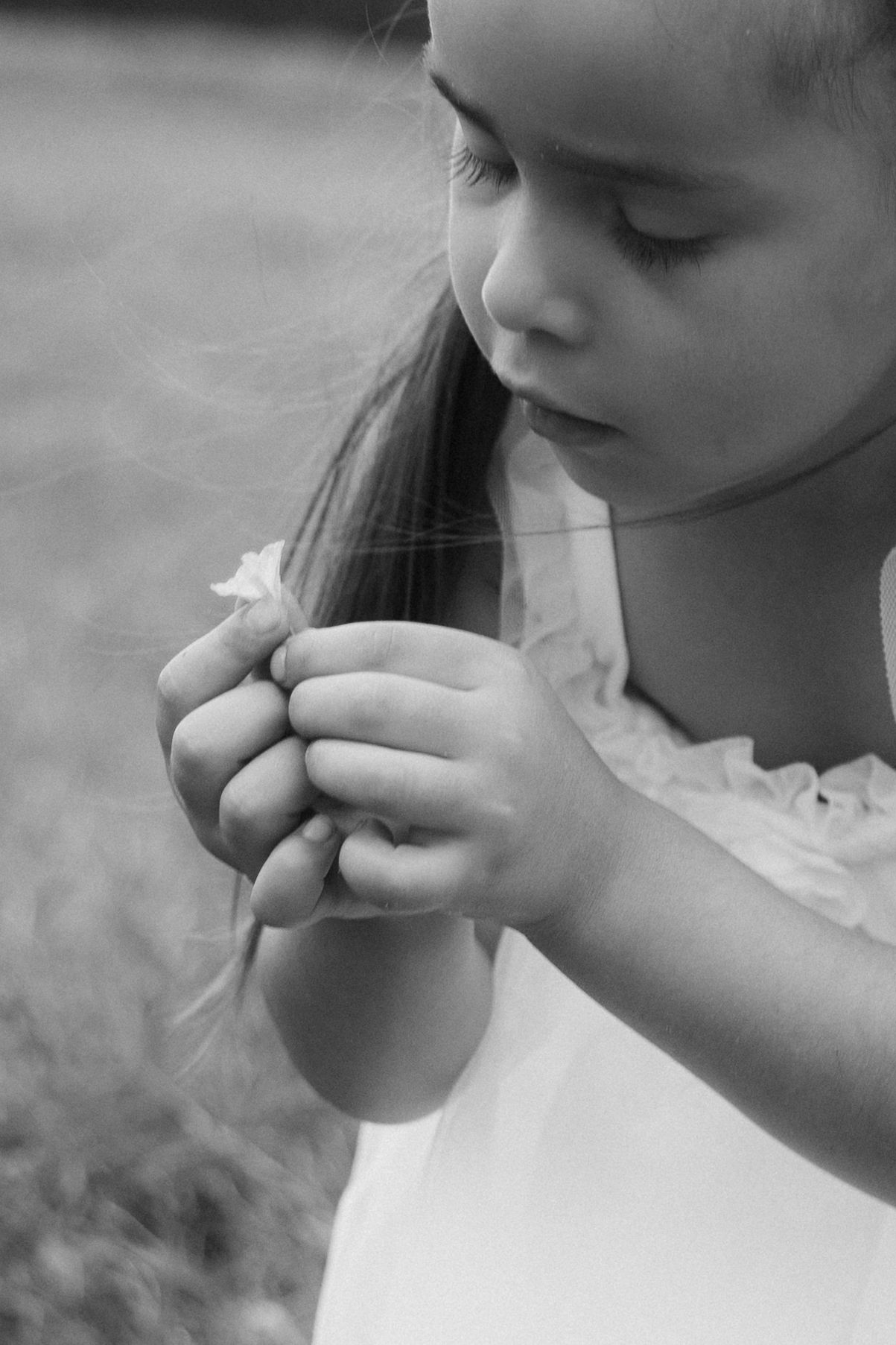 Black and white photo of a young girl in a white dress looking down at a small flower held in her hands outdoors.