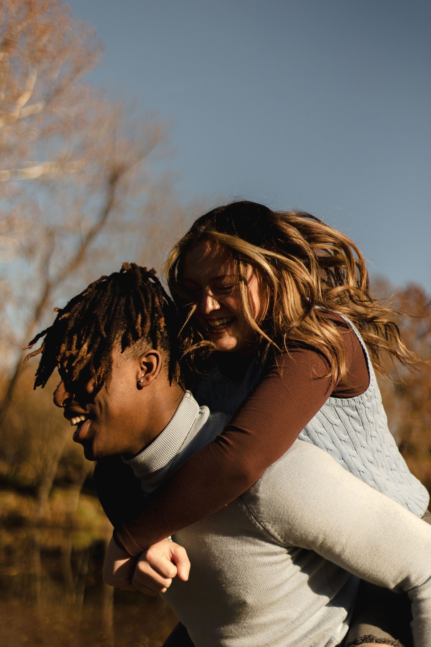 Joyful interracial couple outdoors, man giving woman a piggyback ride against autumn trees and blue sky.