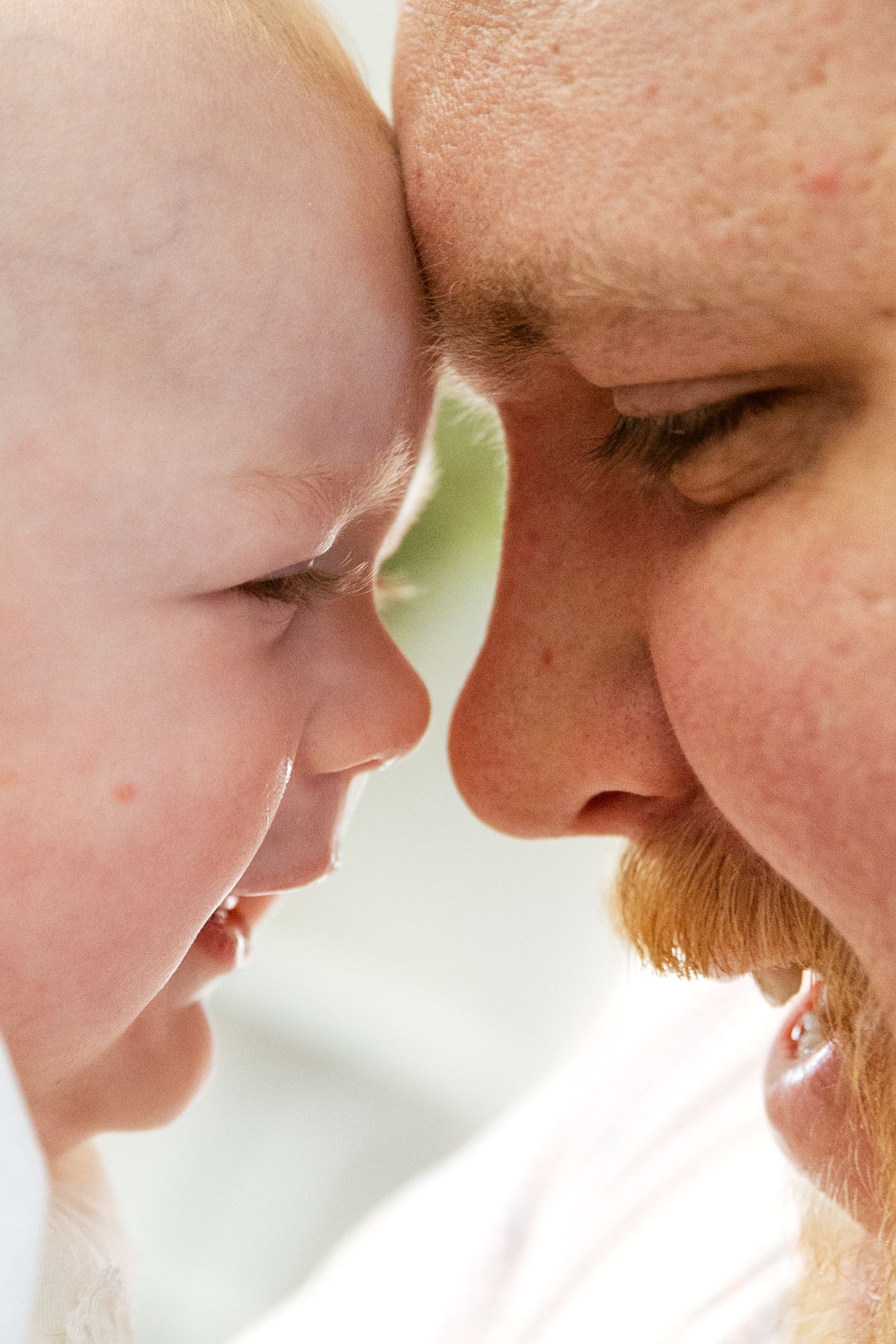 Close-up portrait of a red-bearded father touching foreheads with his smiling newborn baby in a tender moment.