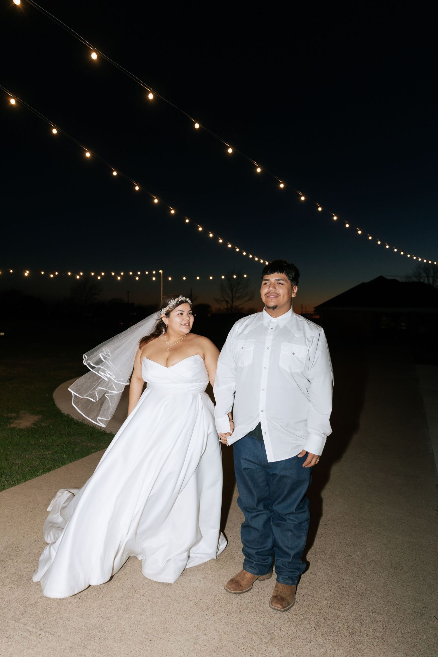 Bride and groom walking hand in hand under string lights on a dark evening path at their wedding.