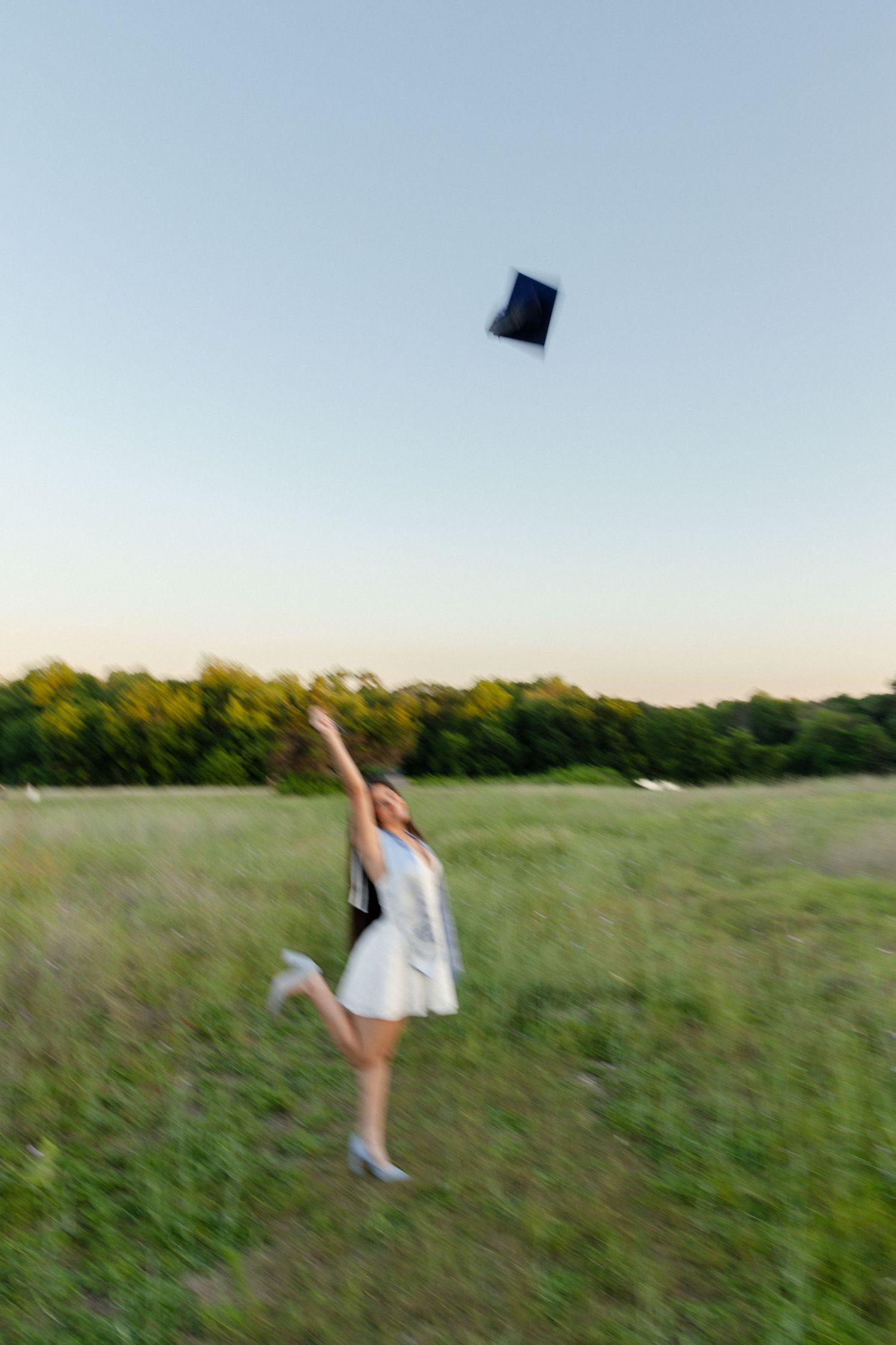 Excited graduate in white dress tossing cap into the air in a green field at dusk.