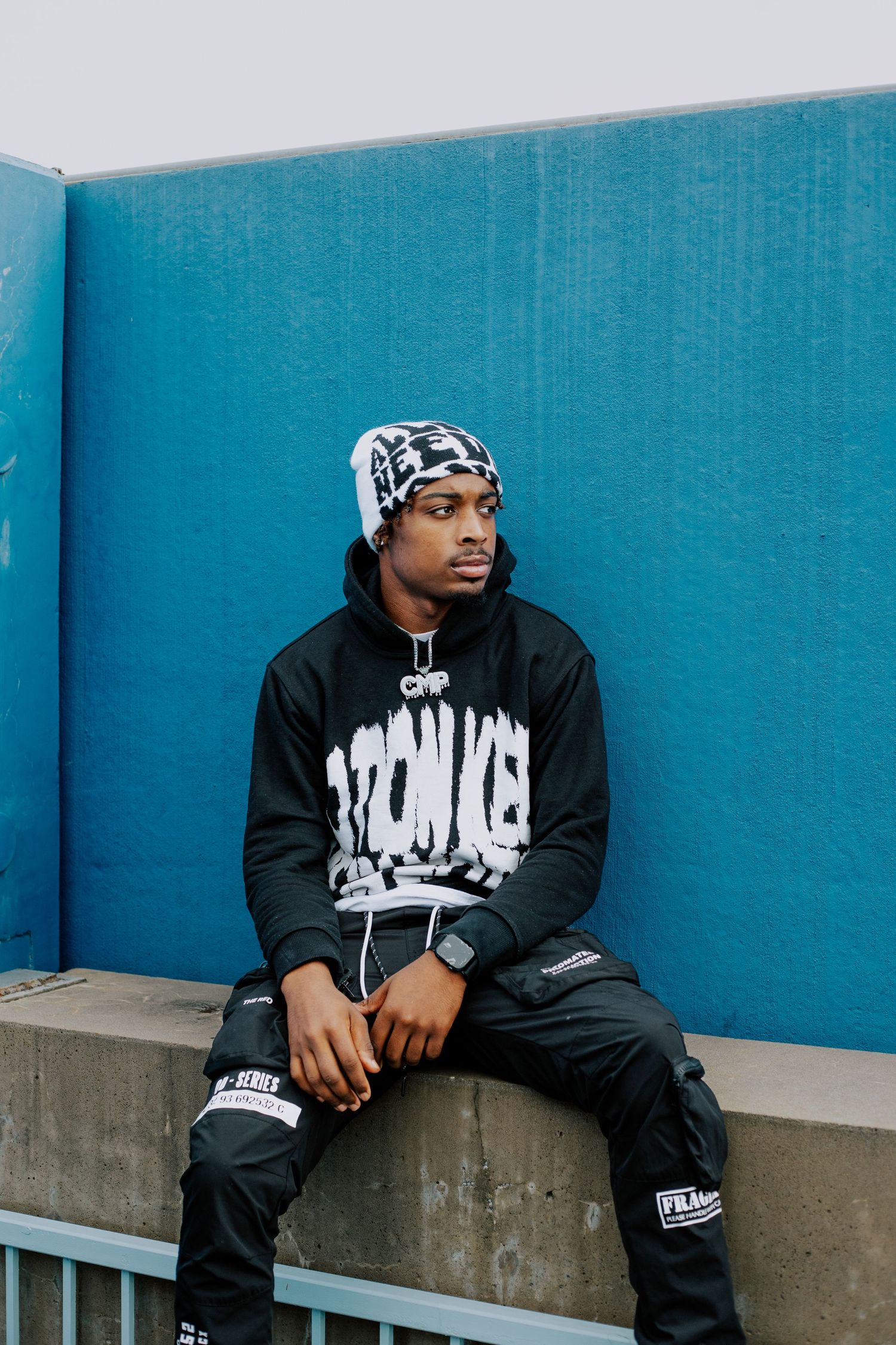 Young Black man in streetwear sitting against a bright blue wall, wearing a hoodie and beanie hat.