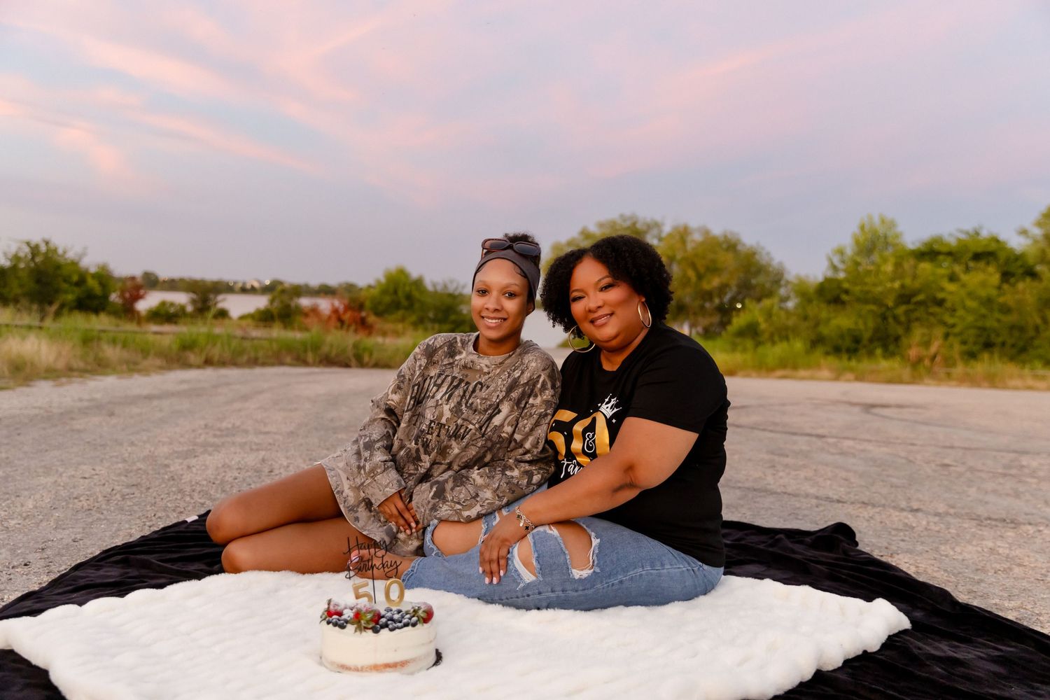 Two women sitting on a black and white blanket outdoors at sunset with a small cake celebrating a birthday.