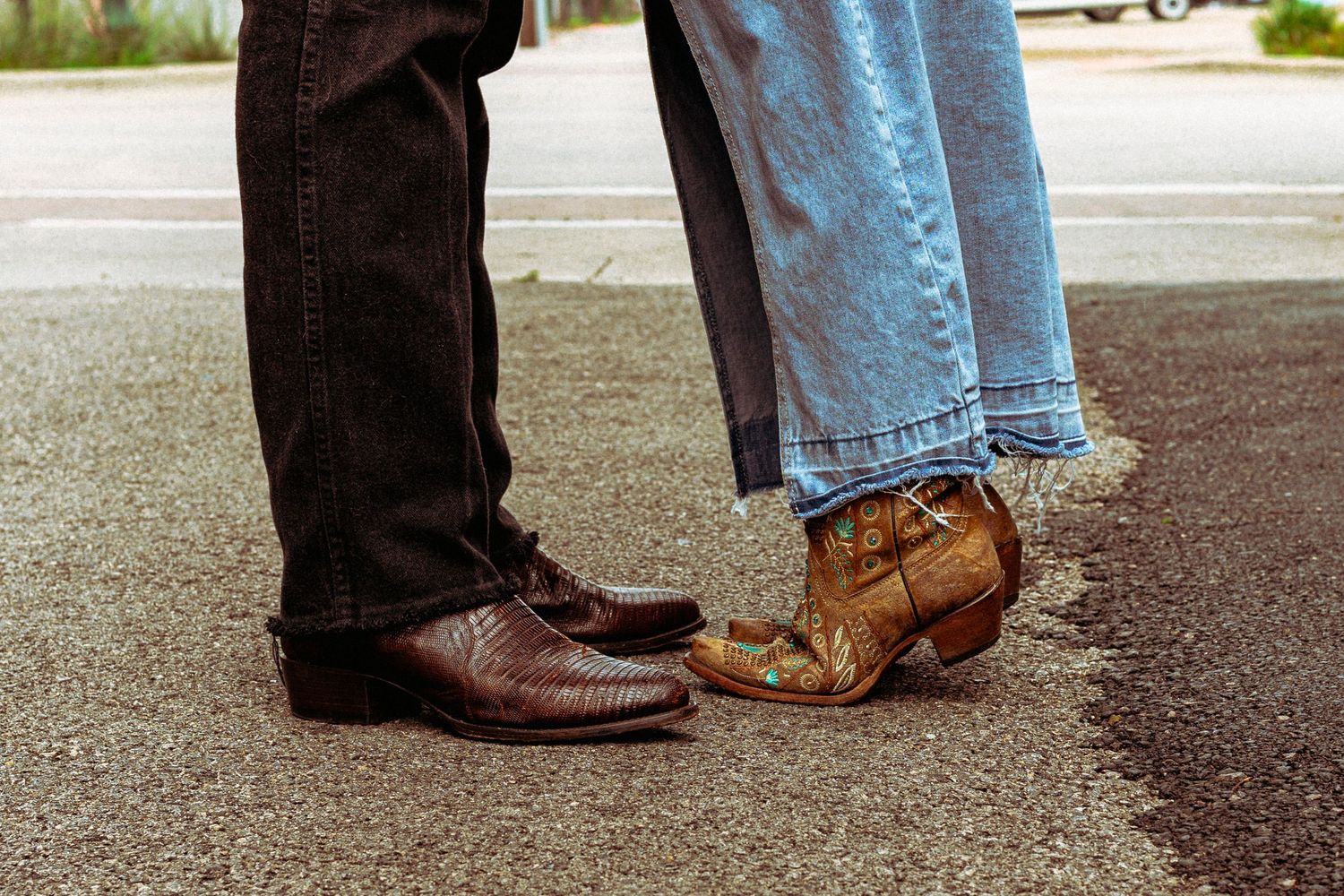 Close-up of two people standing toe-to-toe wearing cowboy boots on asphalt pavement outdoors.