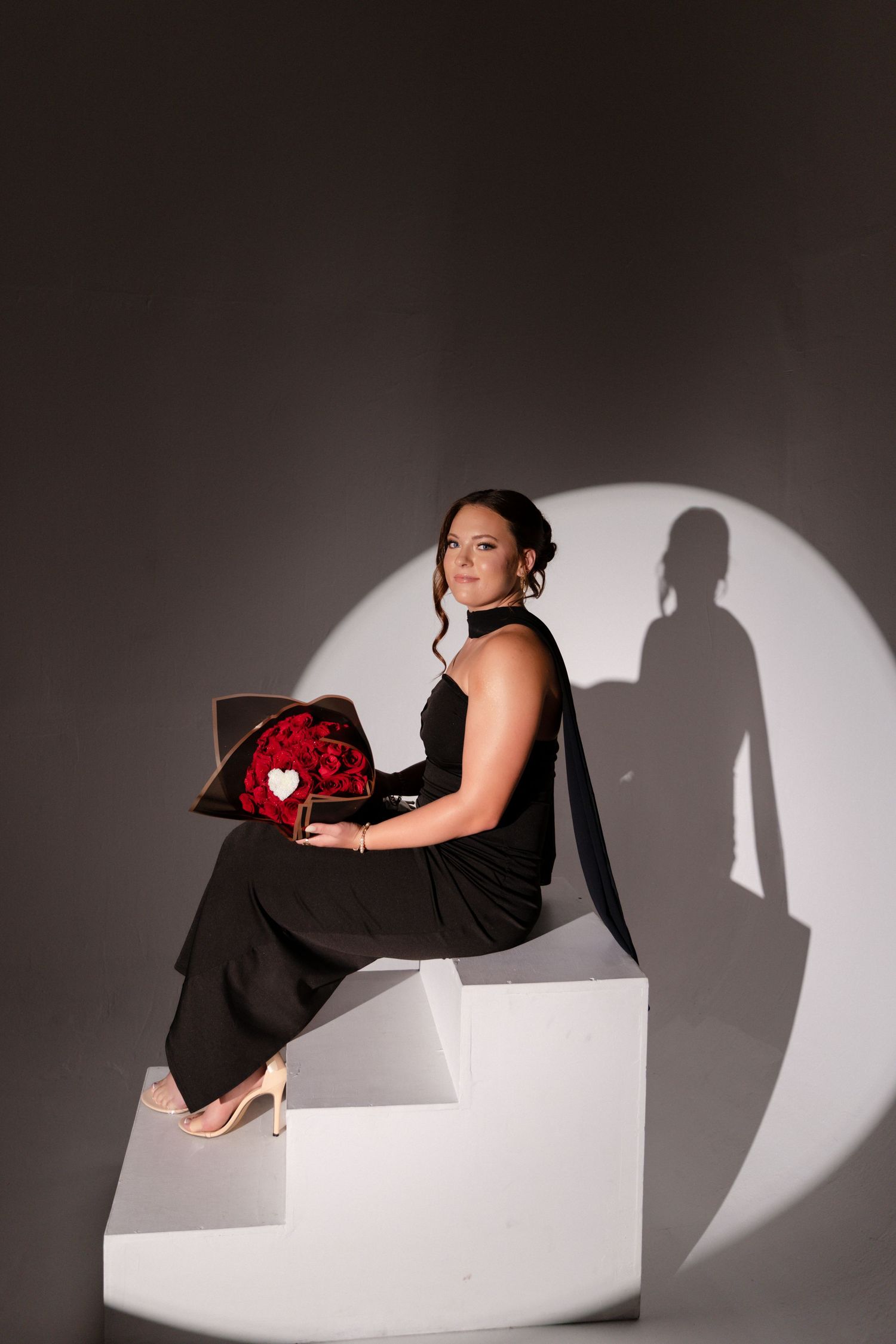 Woman in black jumpsuit sitting on white steps holding heart-shaped rose box, dramatic spotlight shadow backdrop.