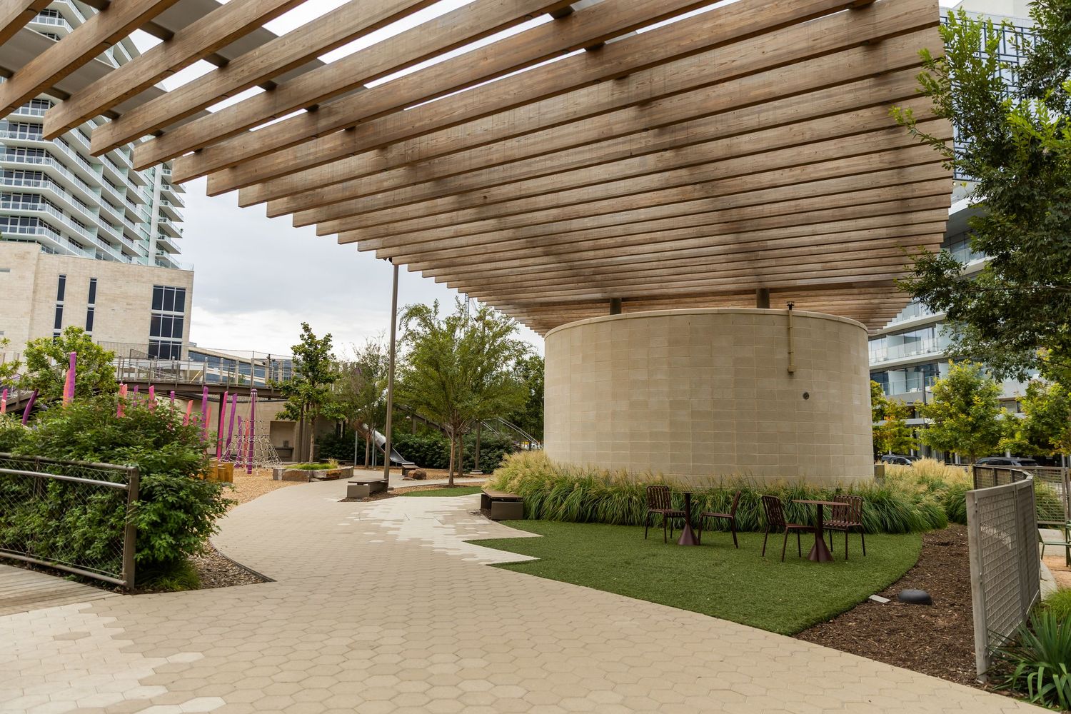 Modern urban park pergola with wooden slat roof, cylindrical concrete structure, lush greenery, and city buildings in background.