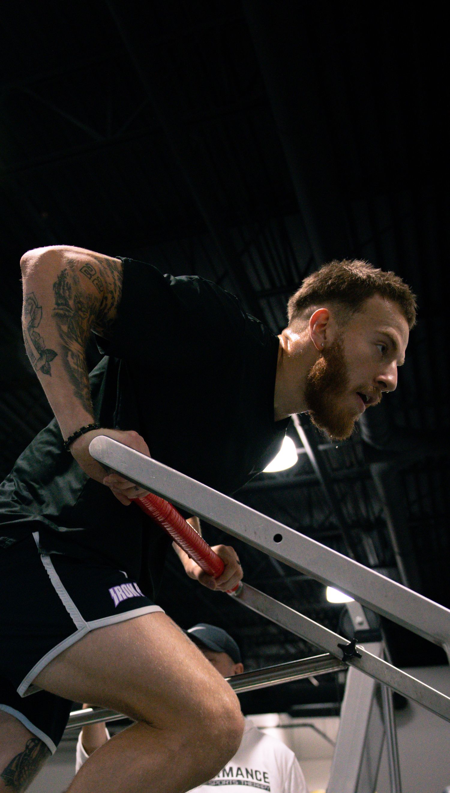 Tattooed male athlete training on parallel bars in dark gym, focused expression during workout session.